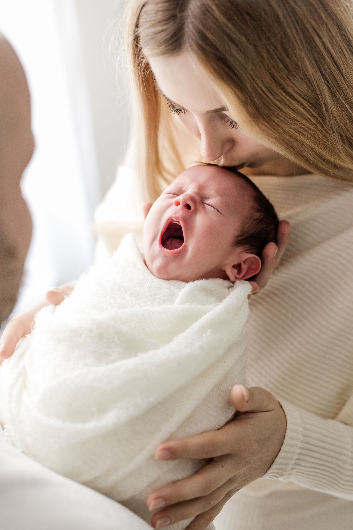 Mother in white sweater kissing crying newborn baby wrapped in soft white blanket.