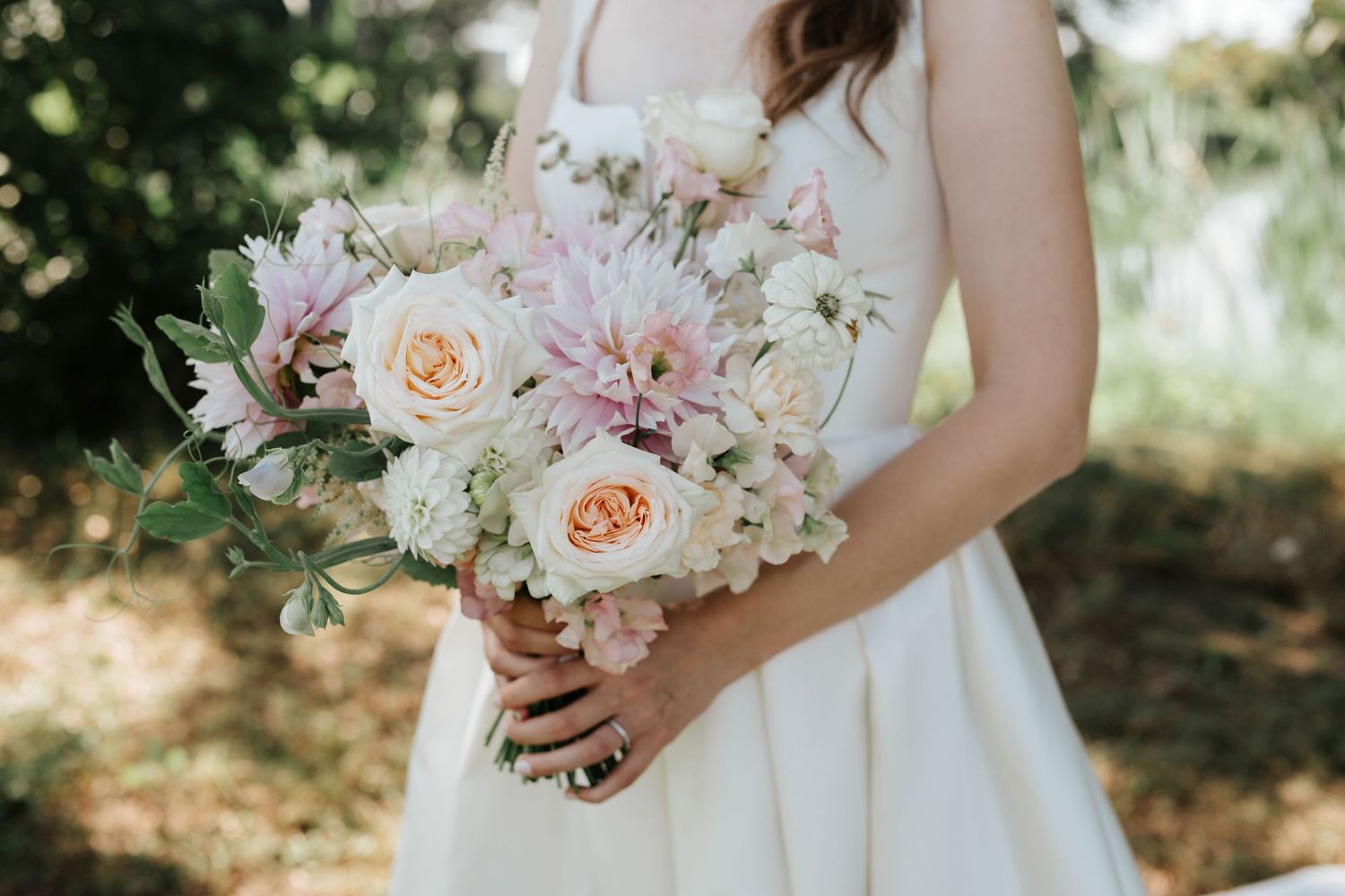 Close-up of bride holding a soft pastel bouquet with roses, dahlias, and greenery in white wedding gown.