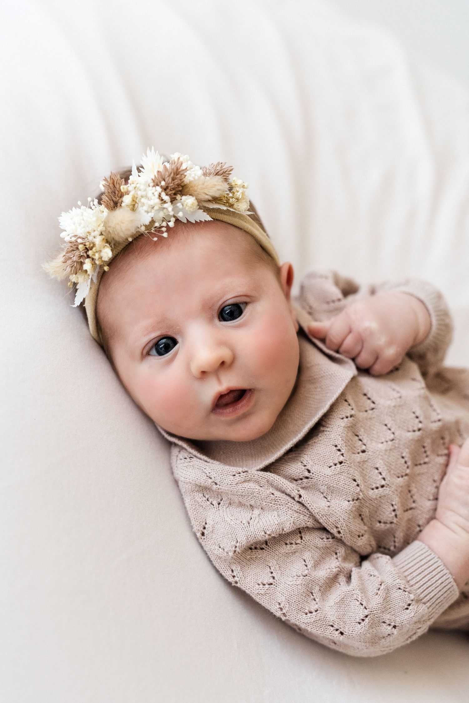 Curious newborn baby girl in a beige knit outfit and dried floral crown, looking up with wide eyes.