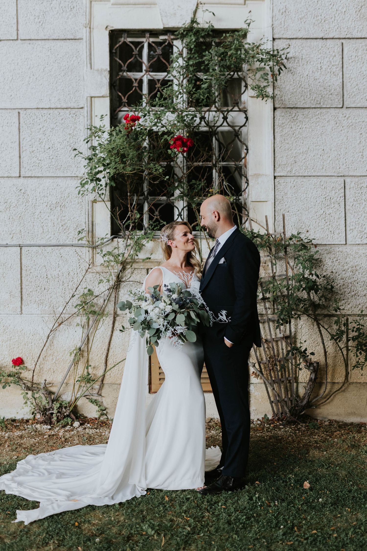 Bride and groom share a romantic moment before a vine-covered castle wall with red roses.