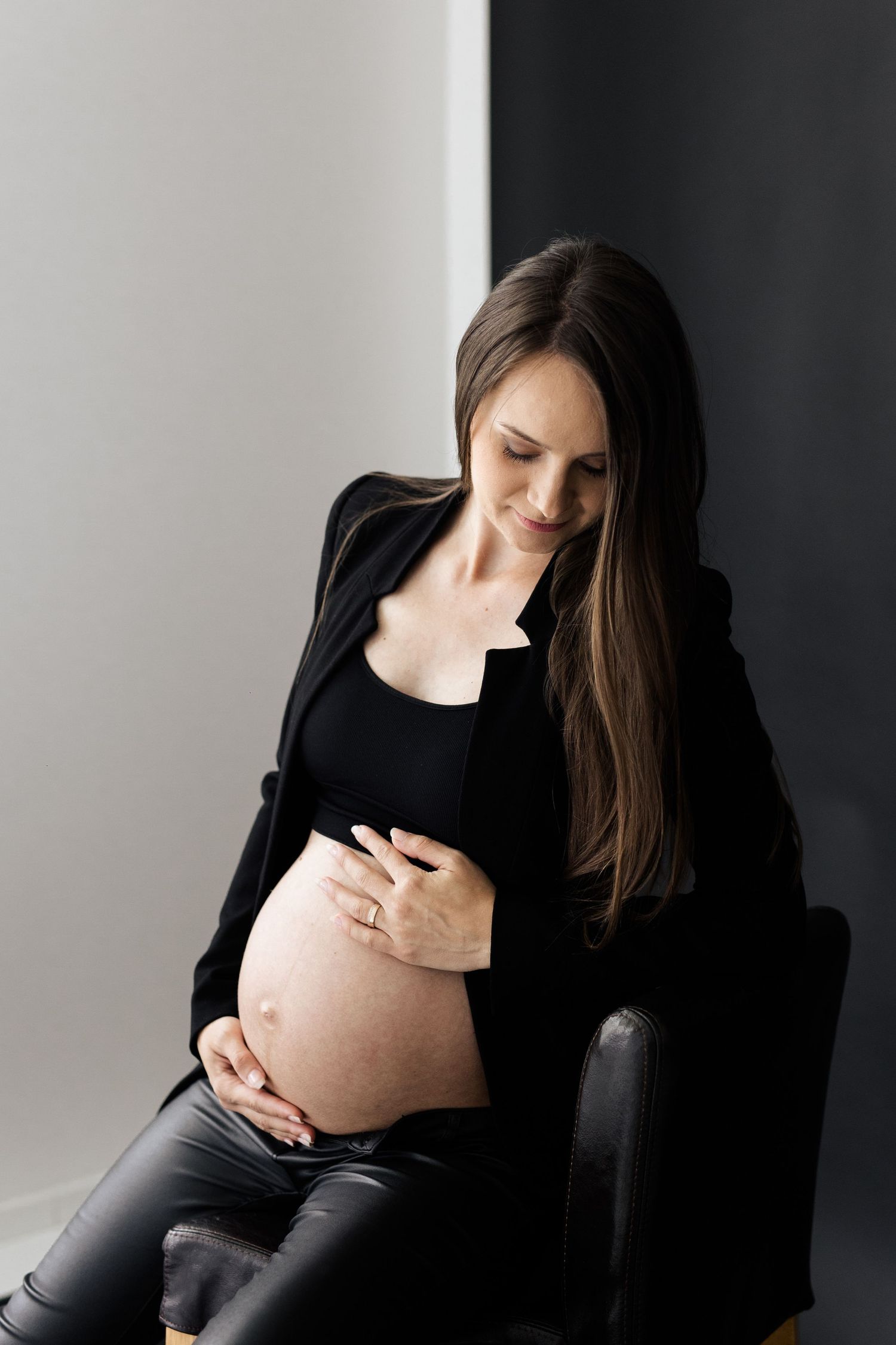 Pregnant woman in black blazer sitting on chair cradling bare bump in studio.