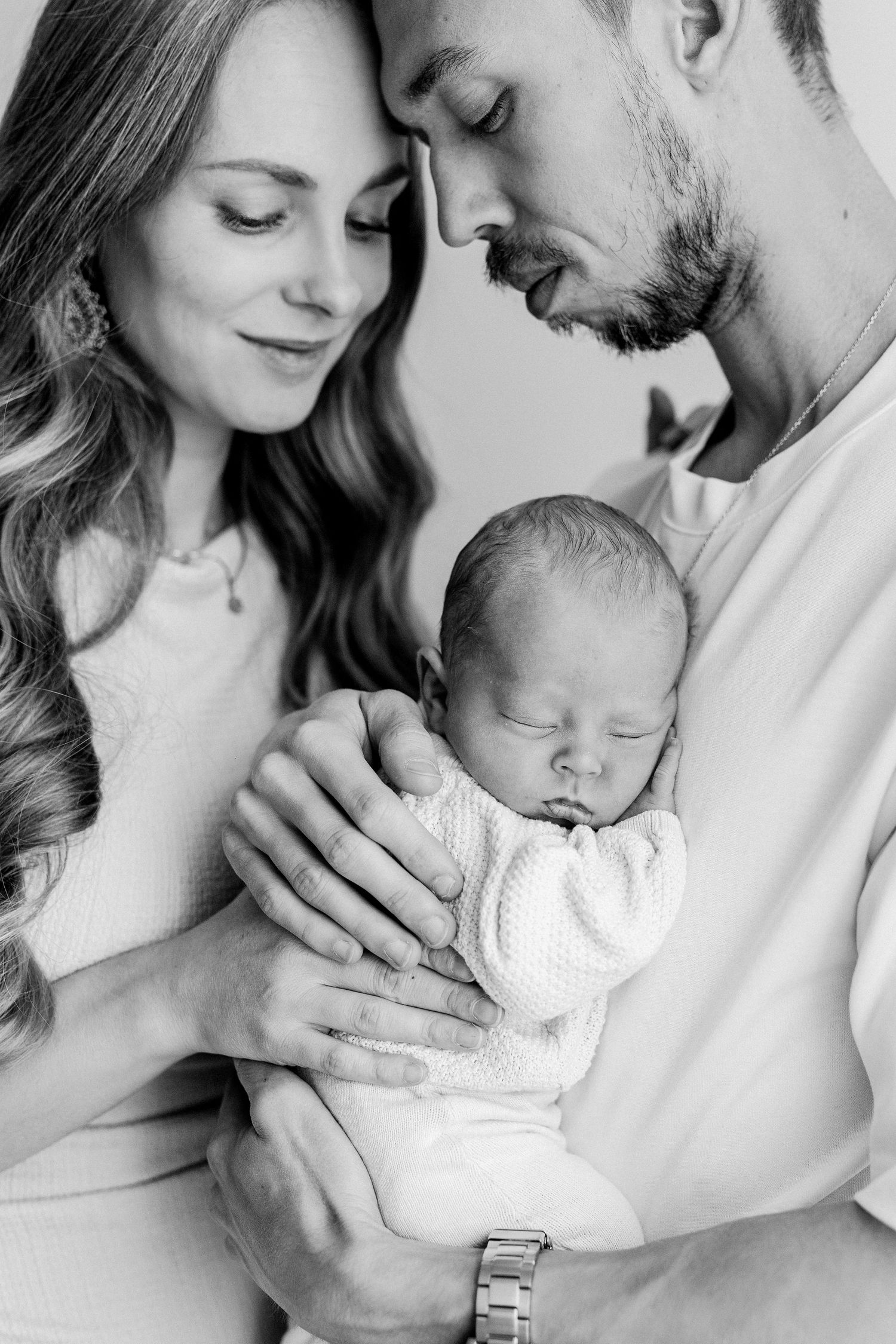 Black and white portrait of parents with foreheads touching while holding sleeping newborn.
