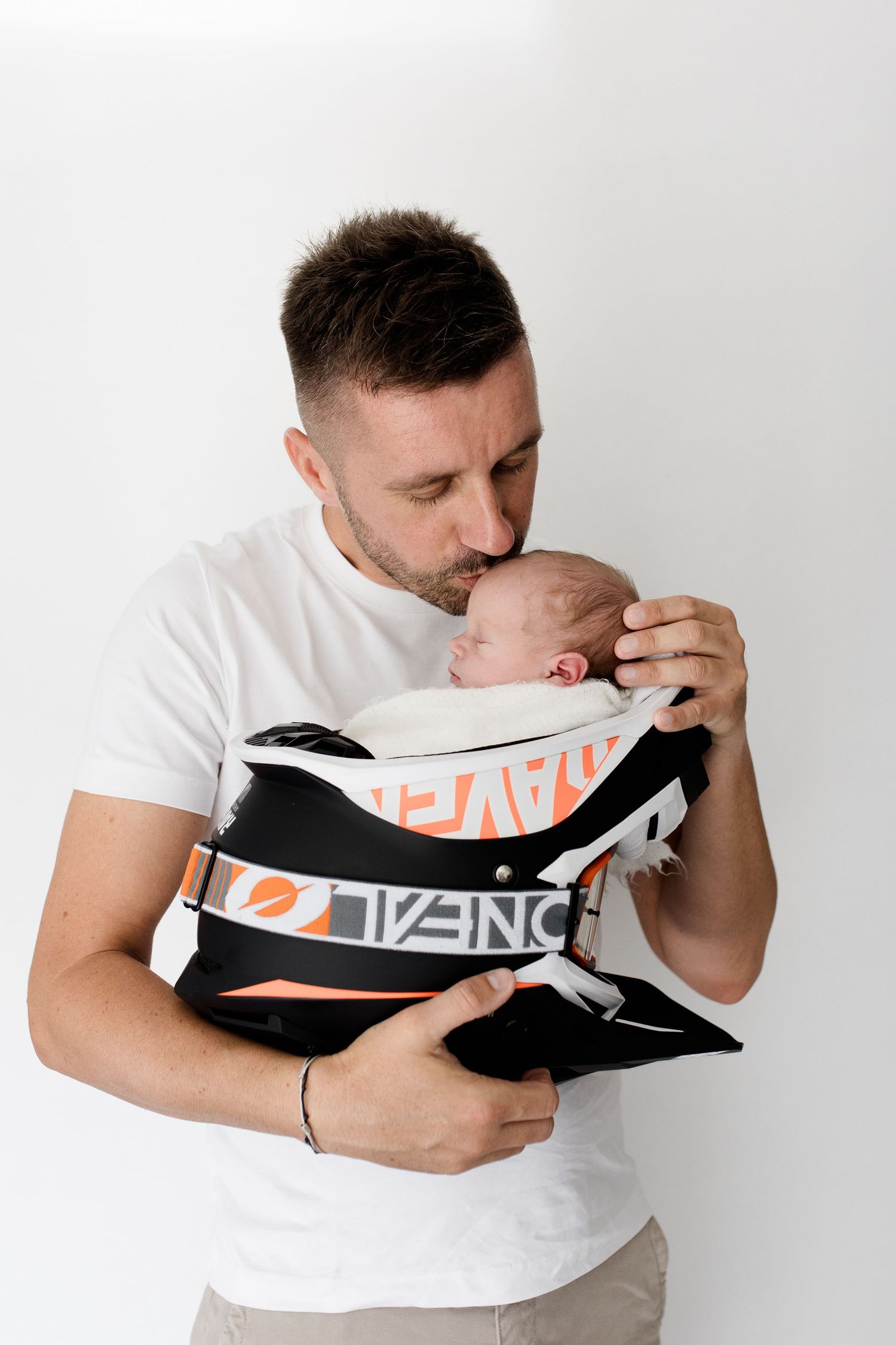 Father in white t-shirt kisses his newborn baby nestled in a black and orange baby carrier.