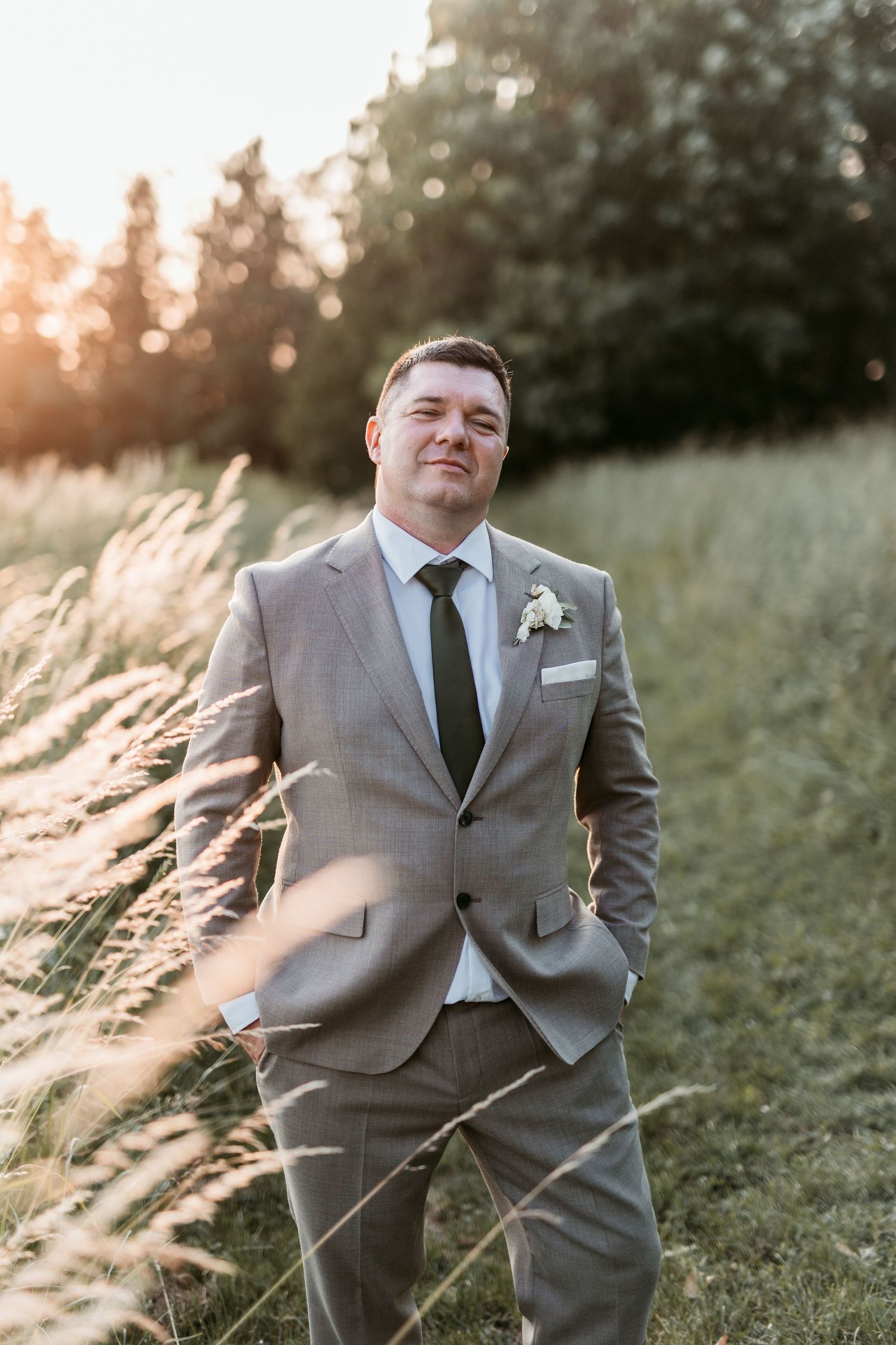 Samozavesten ženin v sivi obleki z naprsnim cvetom, poza na zlato osvetljenem travniku, visoka trava v ozadju.