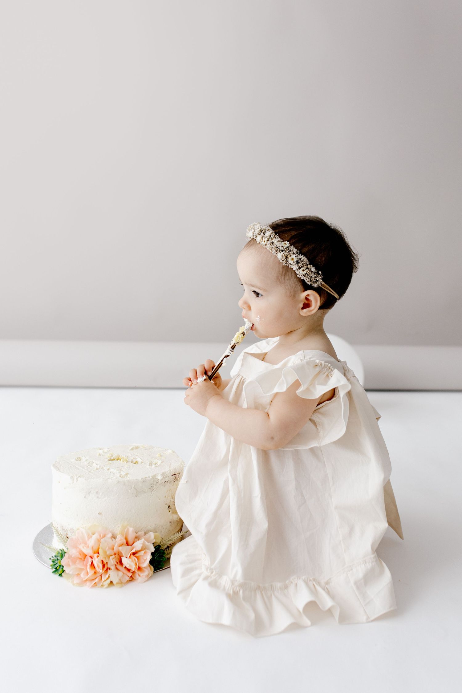 Baby girl in white dress with floral crown licking spoon beside birthday cake.