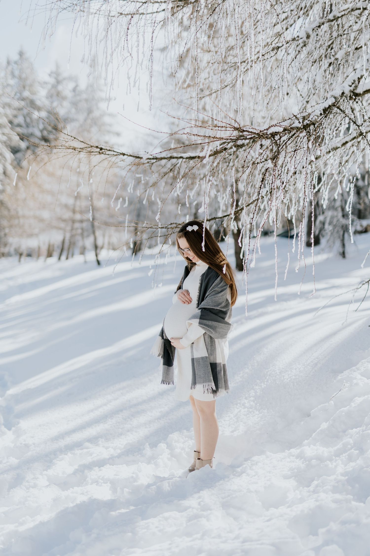 Pregnant woman in white dress and plaid wrap standing in snowy winter forest.