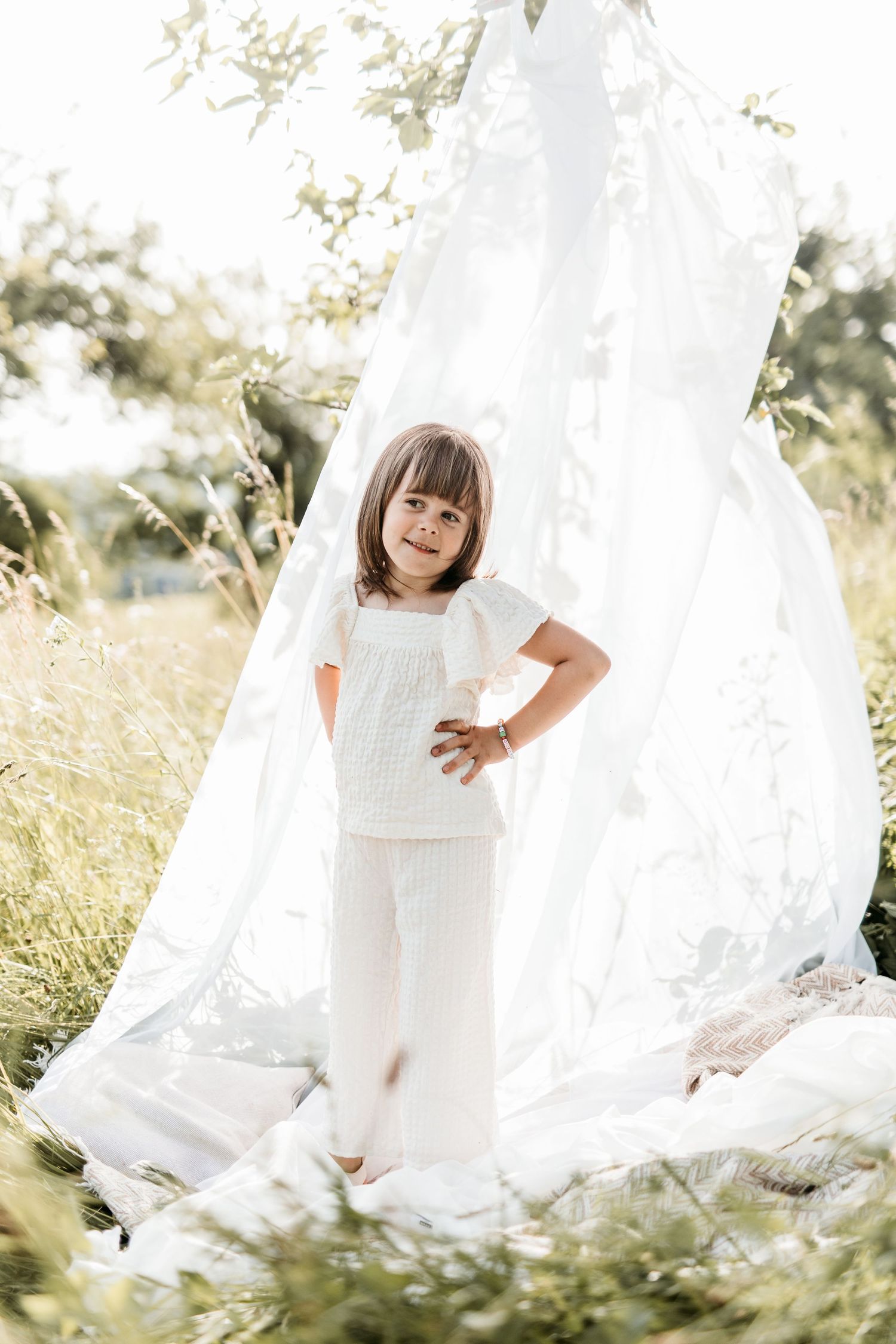 Young girl in white outfit standing in a sheer white teepee tent in a sunny meadow.