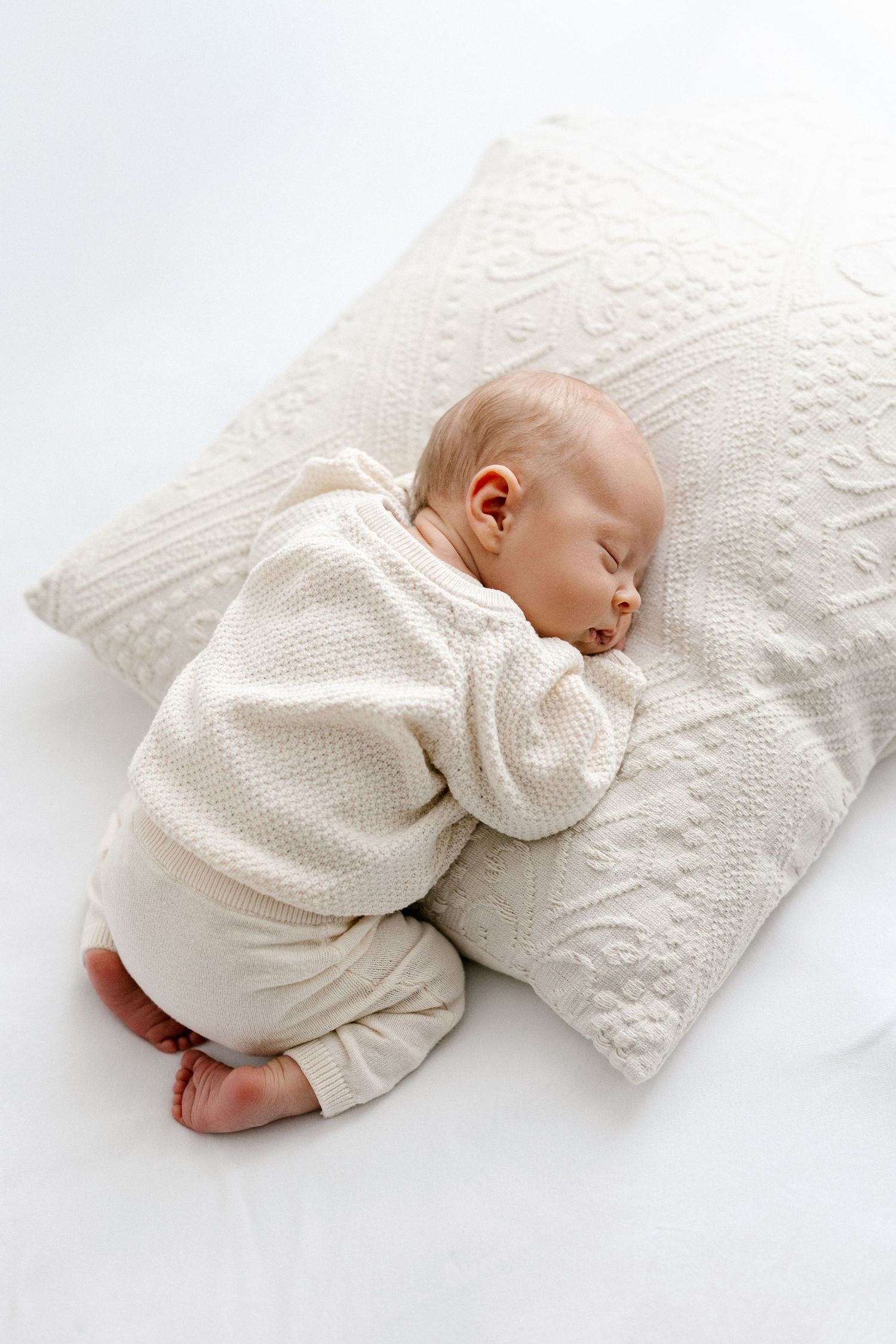 Sleeping newborn baby in white knit outfit resting on decorative white pillow.
