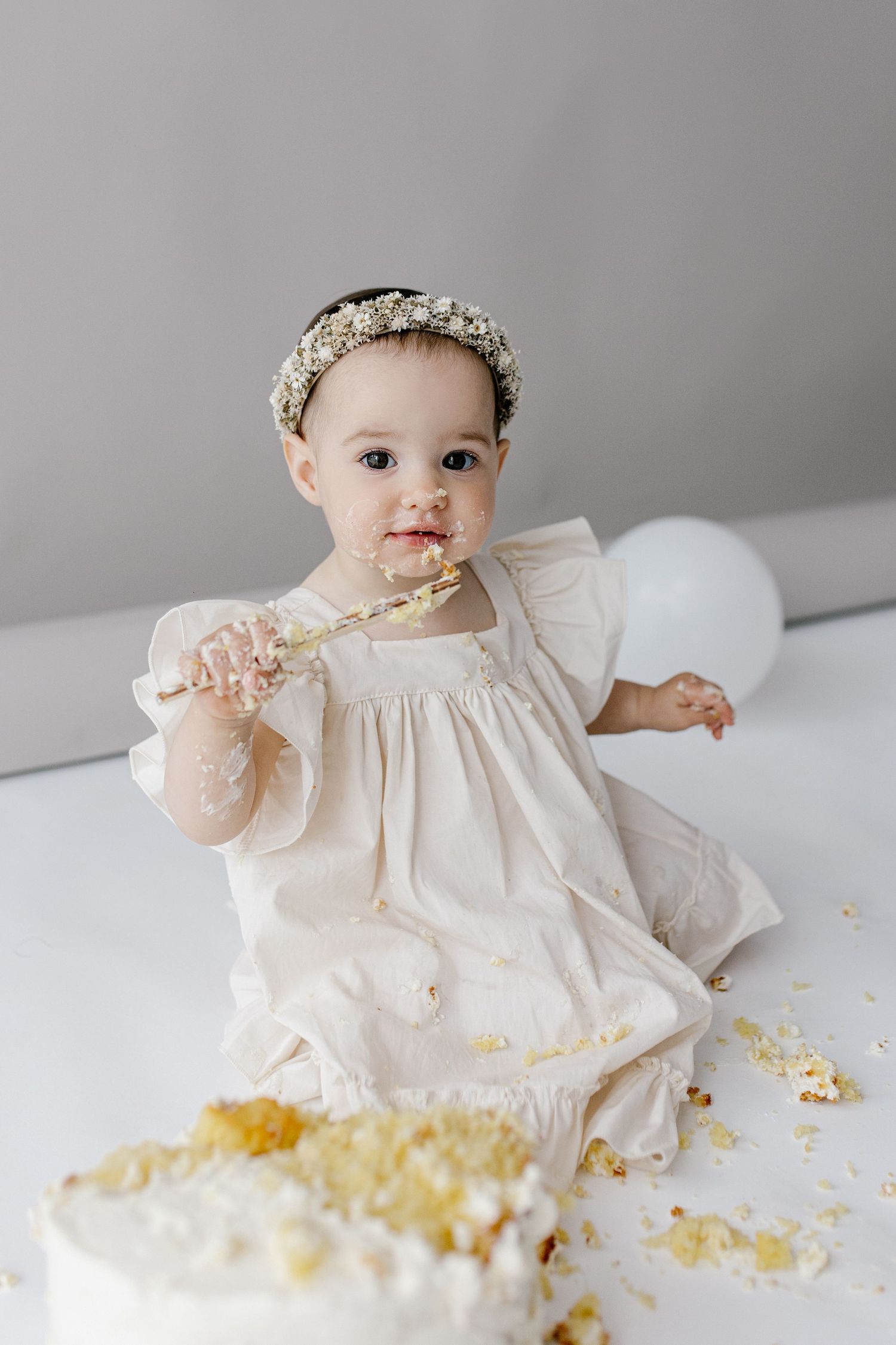 A baby girl in a white dress and floral headband smashes her first birthday cake, holding a fork with frosting.