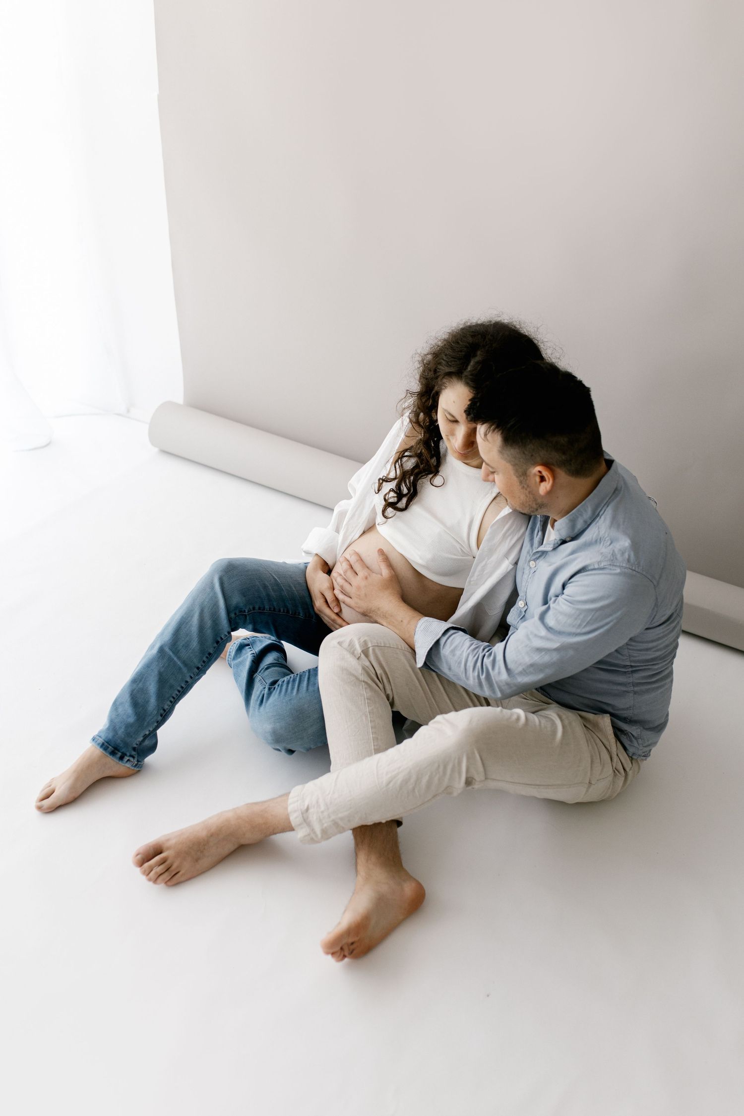 Couple sitting on floor together, man tenderly embracing pregnant woman with baby bump near window.