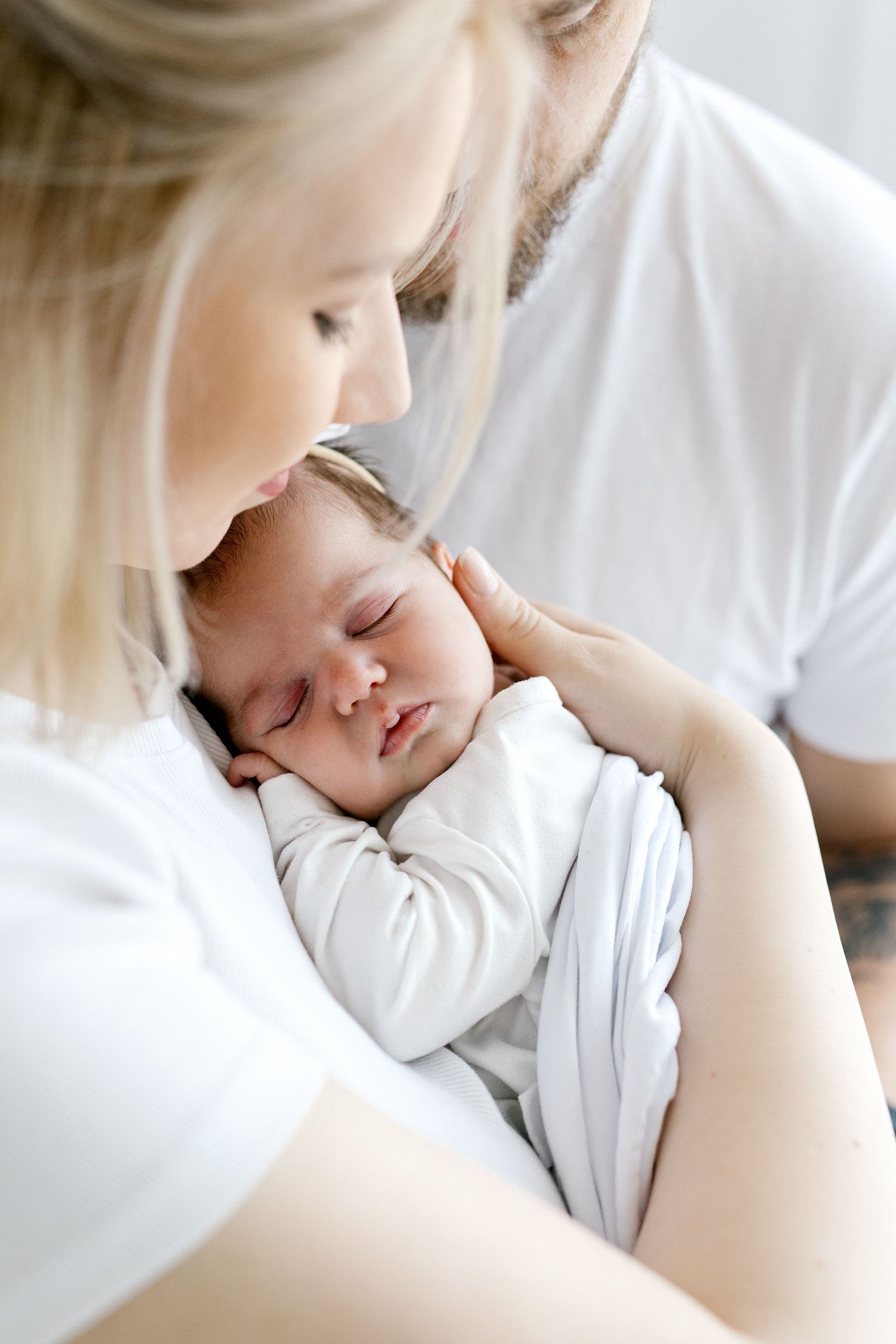 Sleeping newborn baby in white outfit rests peacefully in mother's arms while father looks on lovingly.