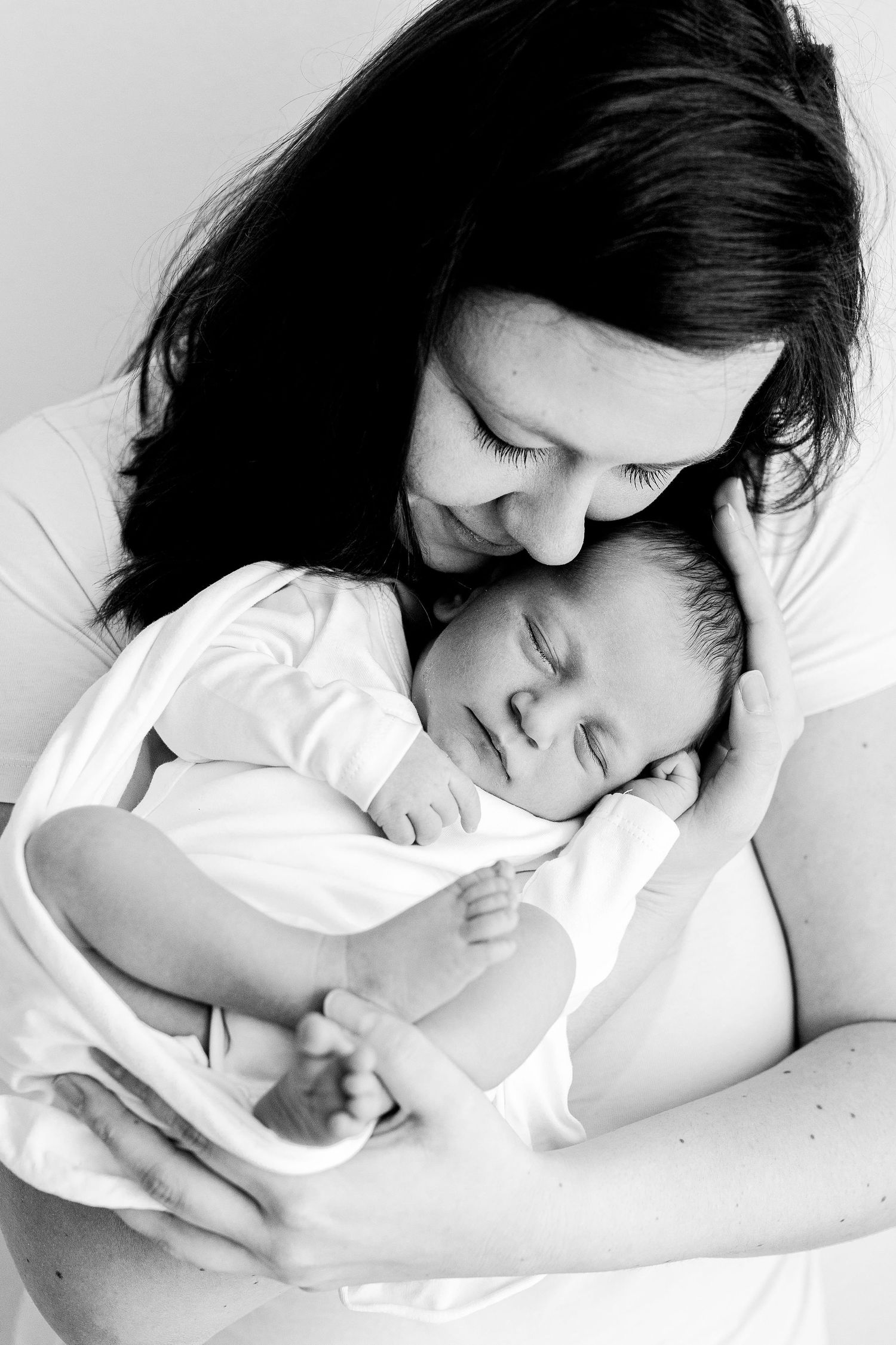 Black and white portrait of dark-haired mother tenderly holding sleeping newborn baby close.