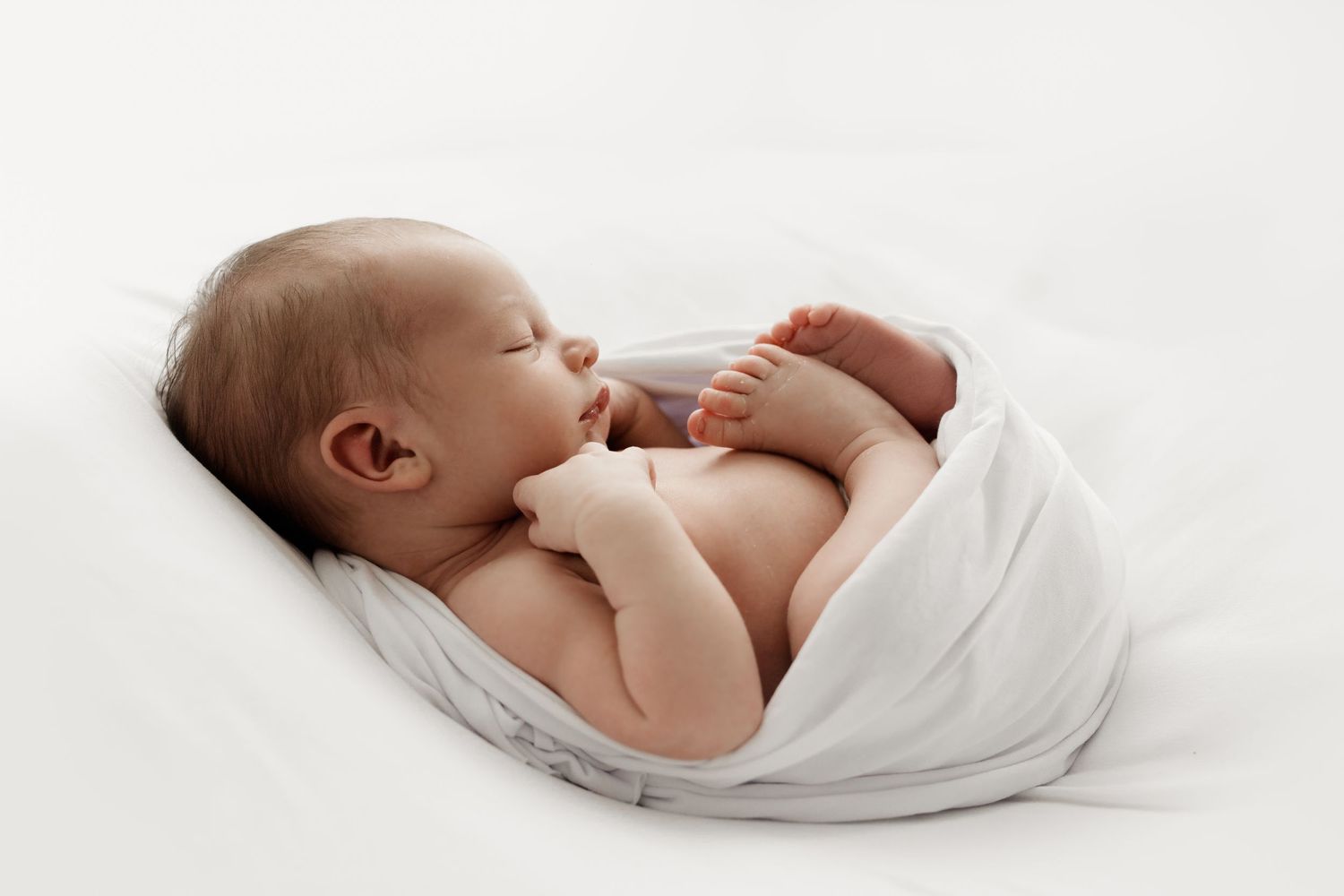 Serene newborn baby sleeping on side wrapped in a soft white swaddle blanket.
