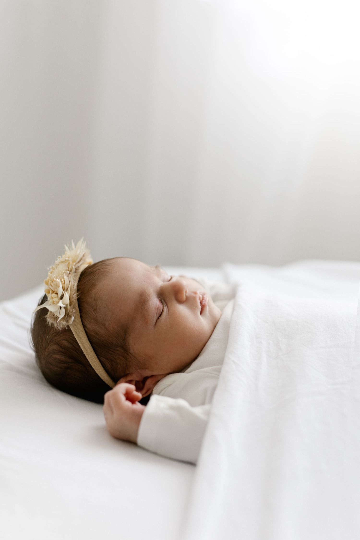 Peaceful newborn baby girl wearing a floral crown sleeps on a white blanket in soft natural light.