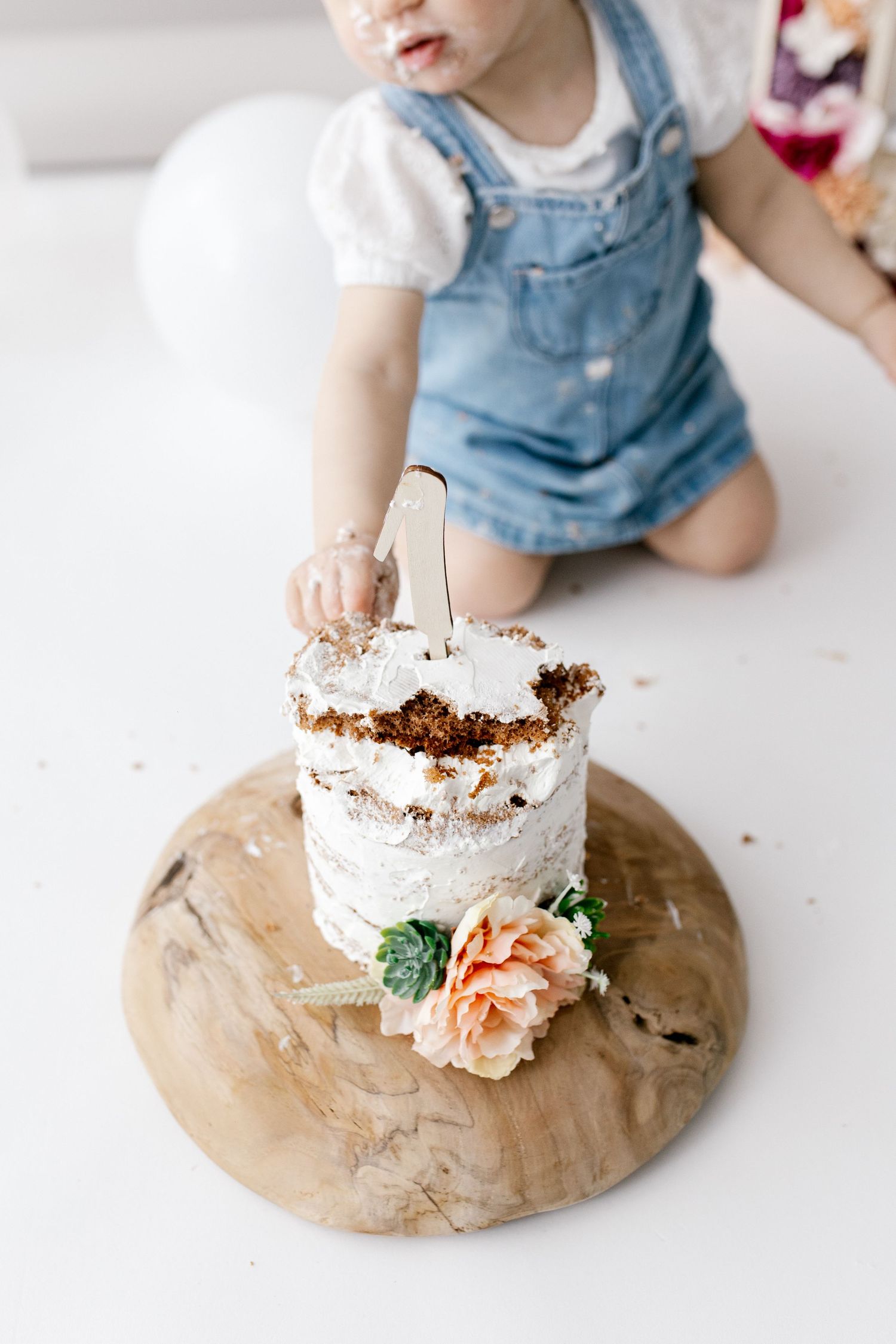 Baby in denim overalls reaching into smash cake on wooden board with flower.