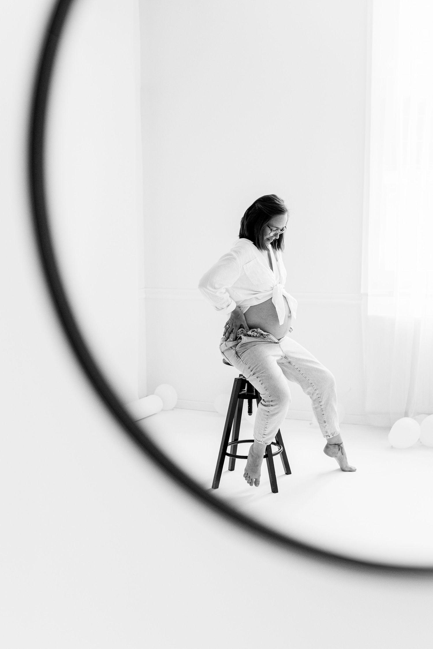 Black and white reflection of pregnant woman sitting on stool viewed through round mirror.