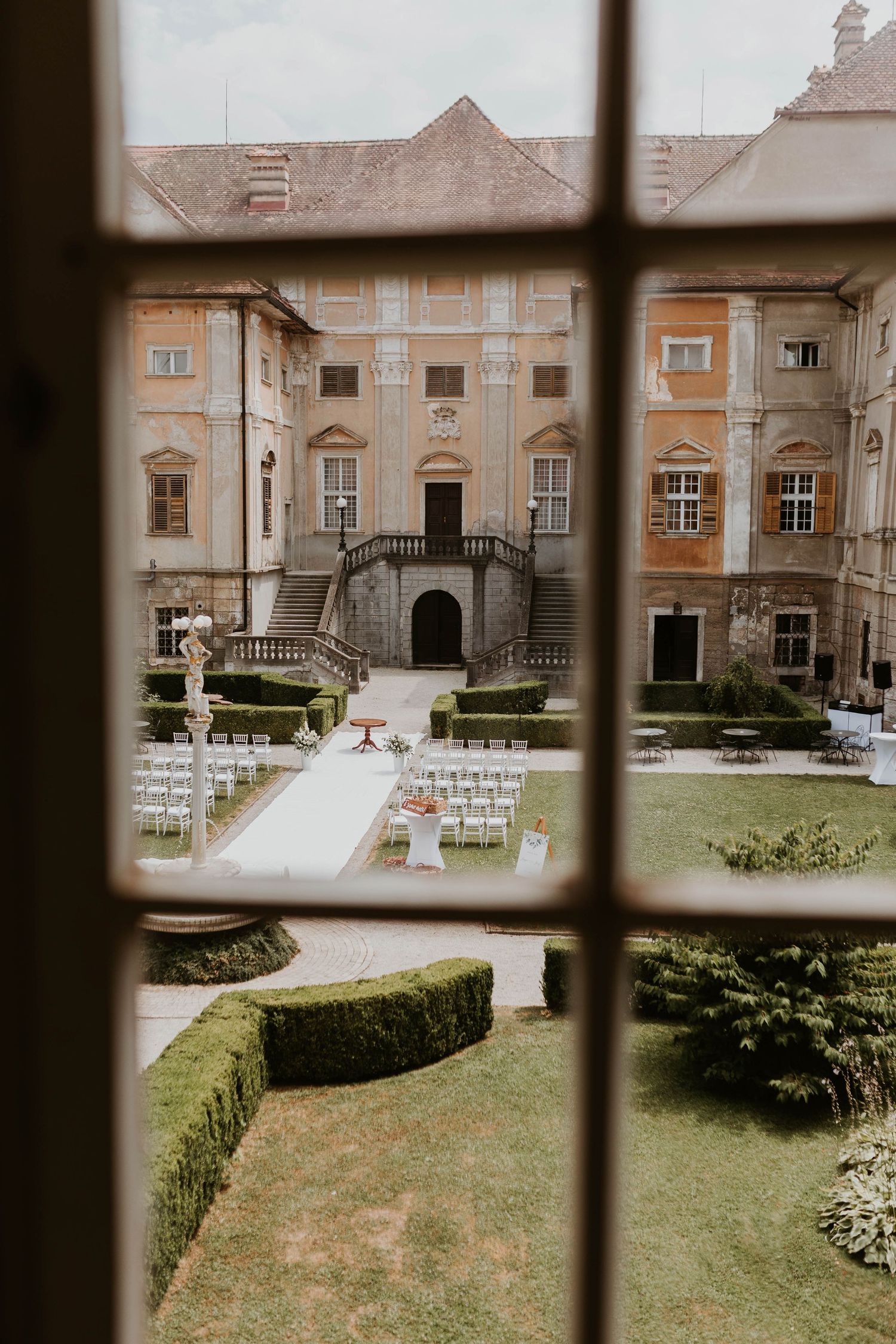Elegant castle courtyard wedding ceremony setup with white chairs and manicured hedges viewed through a window.