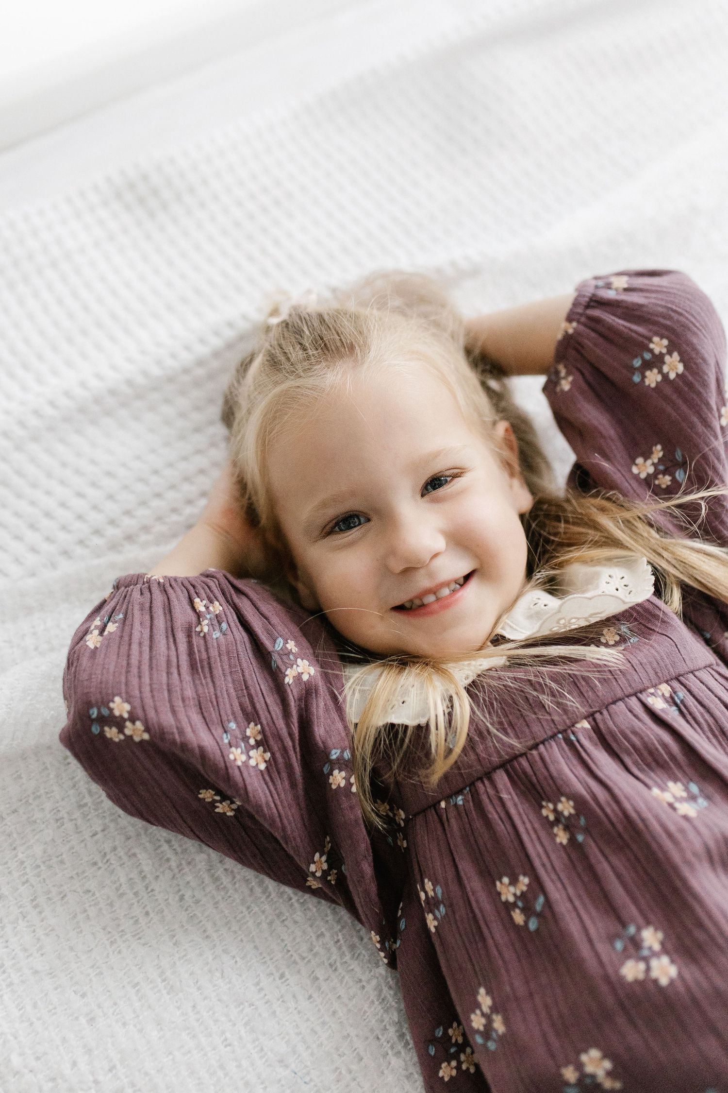 Smiling young girl in purple floral dress lying on her back on a white blanket.