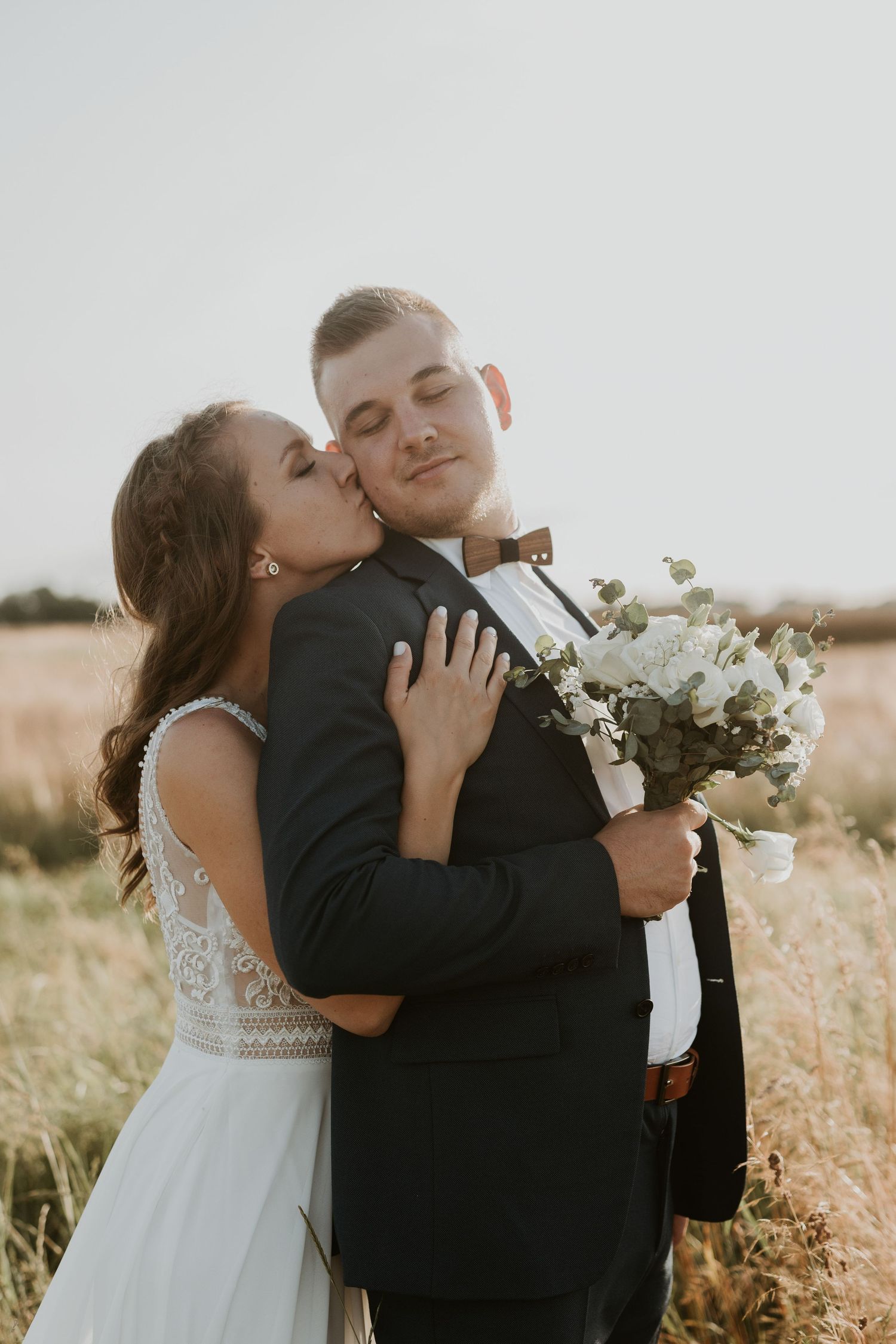 Bride kisses groom on the cheek in a golden field, groom holding a white floral bouquet.