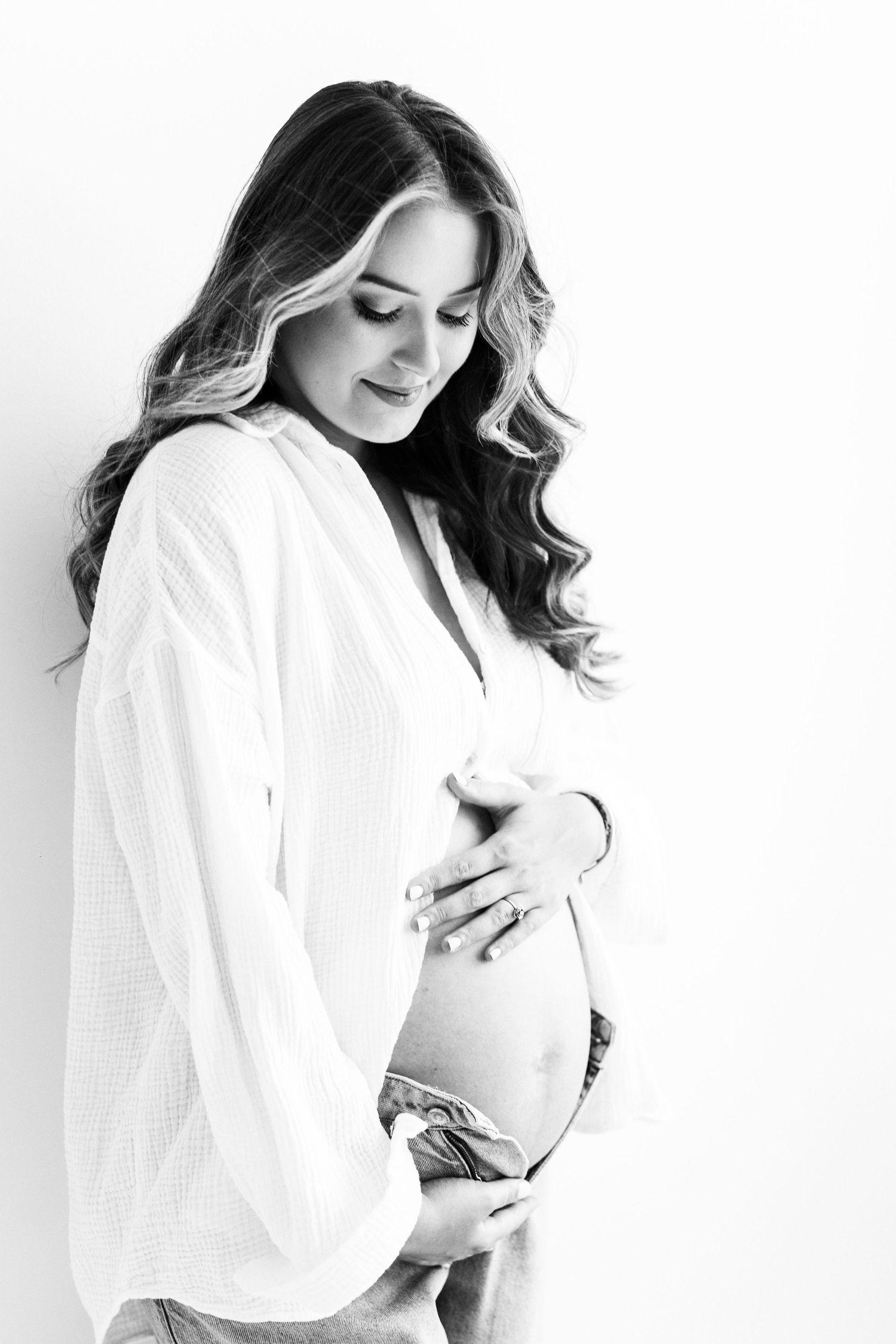 Black and white portrait of beautiful pregnant woman cradling bare baby bump in white shirt.