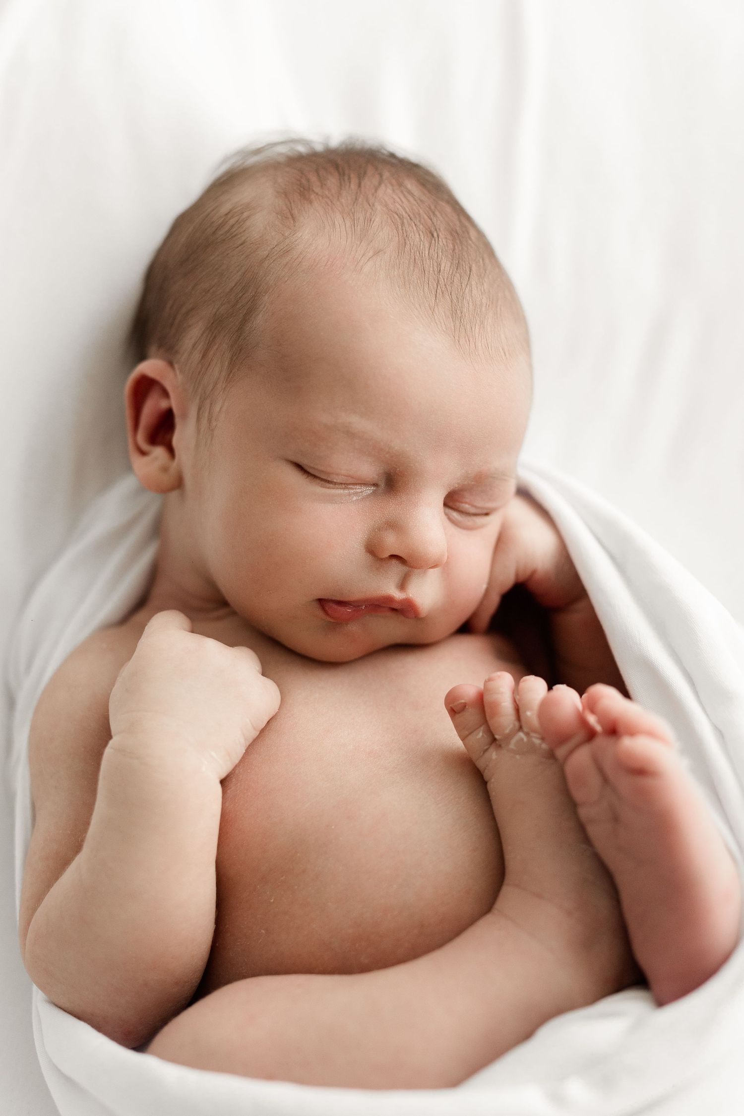 Close-up of a sleeping newborn baby wrapped in white swaddle with tiny hands and feet visible.