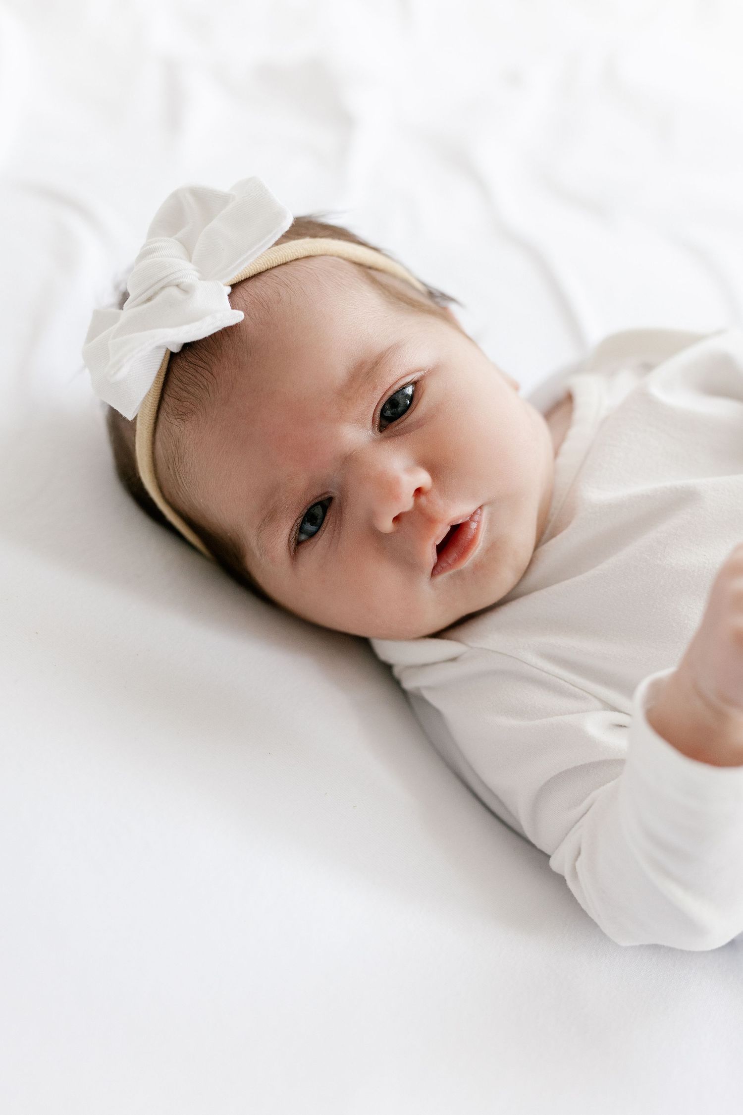 Adorable newborn baby girl in white onesie and bow headband lying on white bedding.