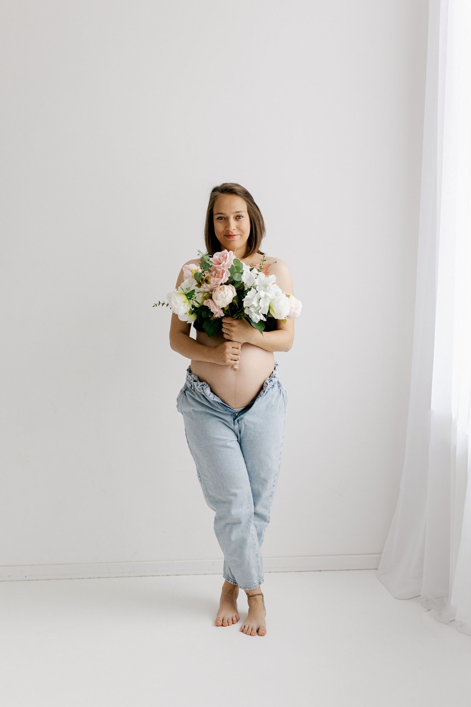 Pregnant woman in jeans holding large bouquet of pink and white flowers covering her bare belly.