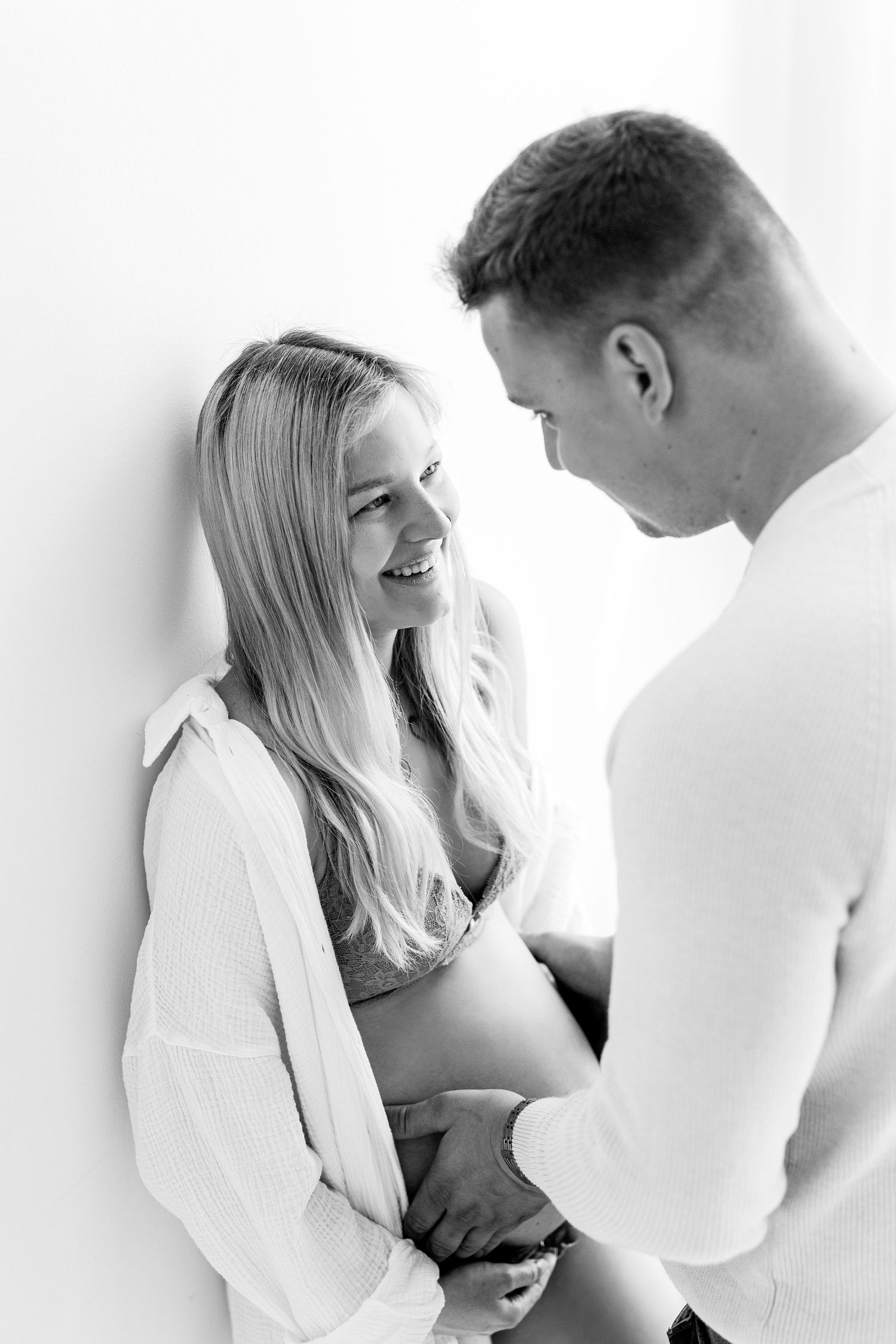Black and white maternity photo of smiling blonde woman with partner holding her pregnant belly.