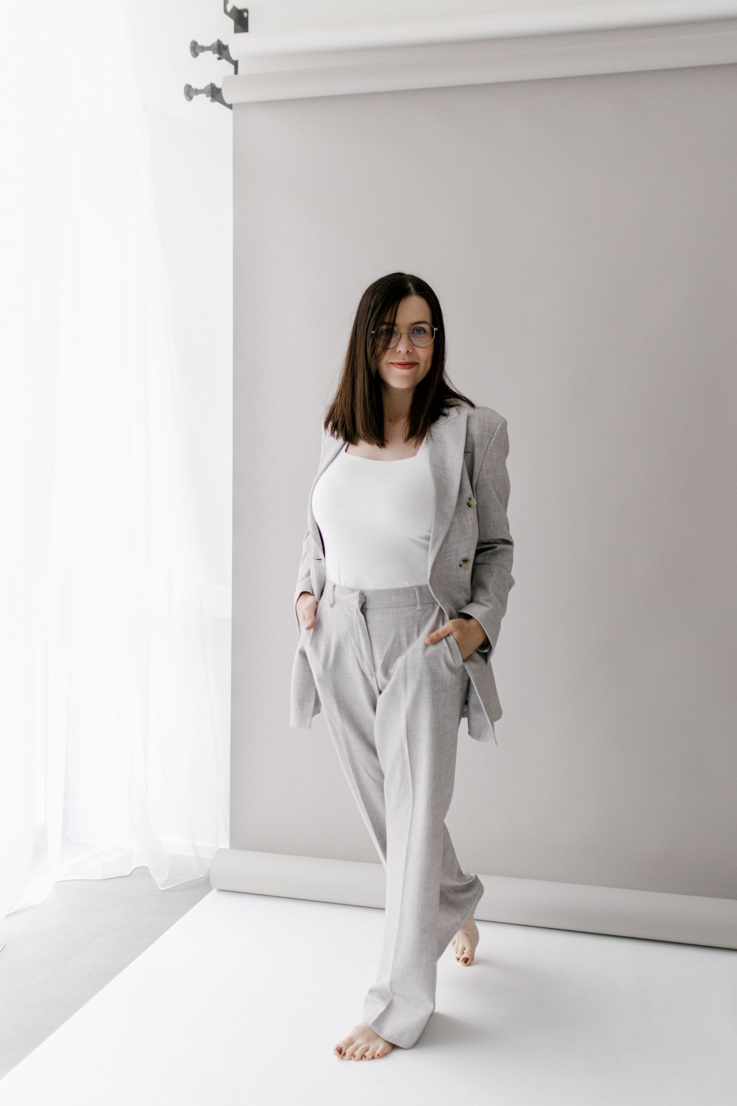 A woman in a gray blazer and trousers poses barefoot in a bright white studio space.