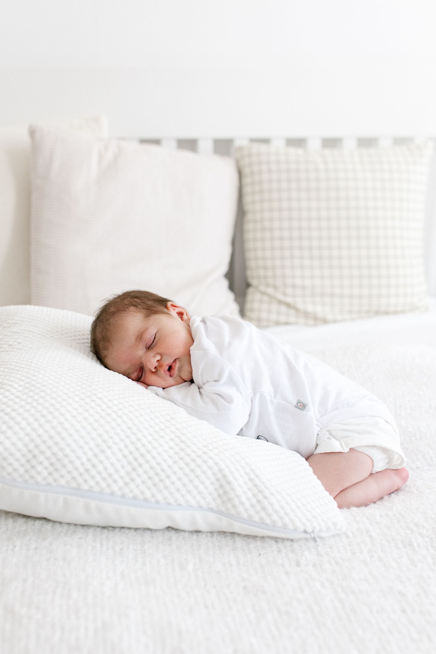 Smiling newborn baby in white outfit lying on a large white pillow on a bed.