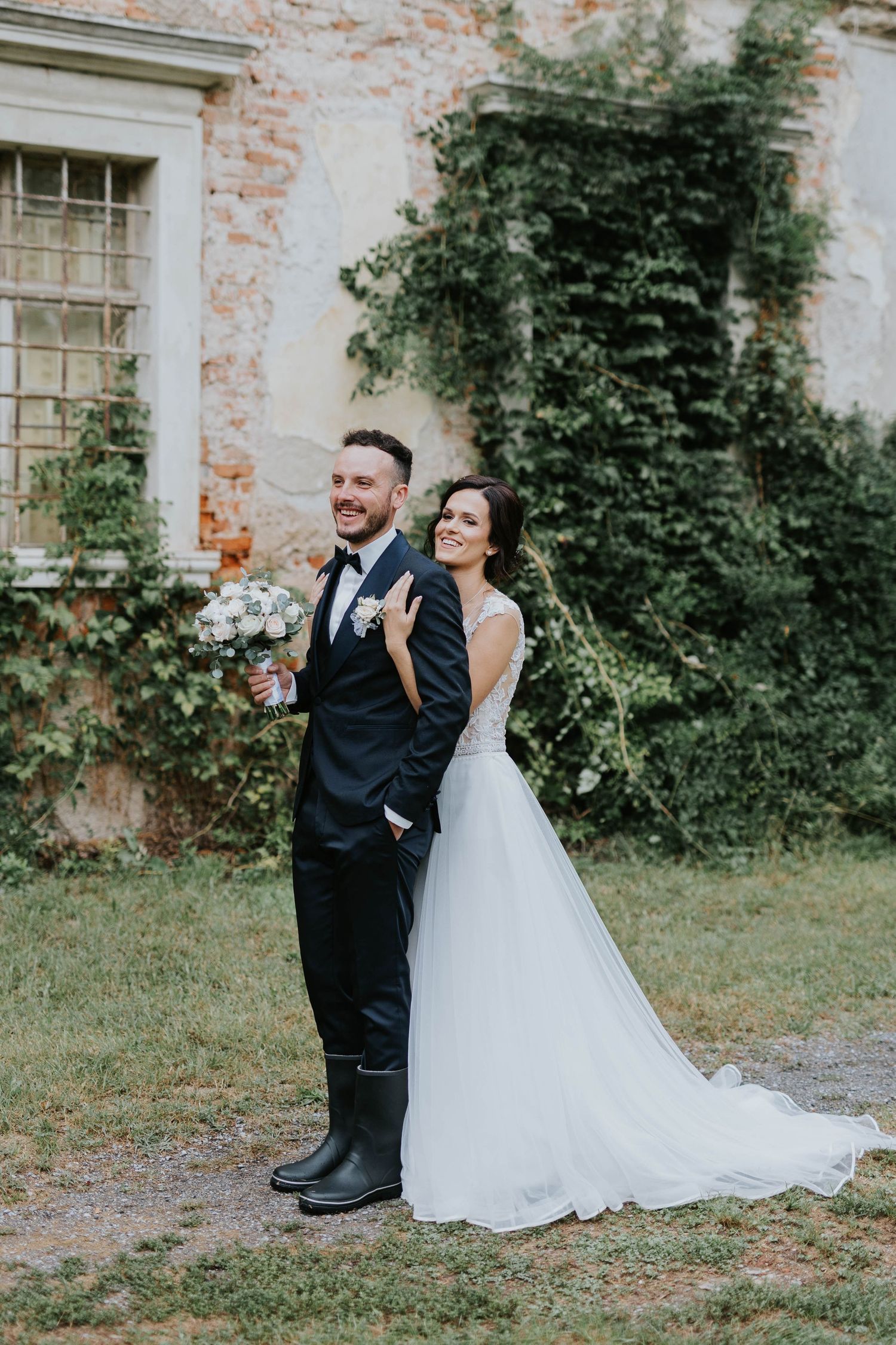 Bride and groom posing outdoors before an ivy-covered brick wall, groom in black tuxedo holding bouquet.