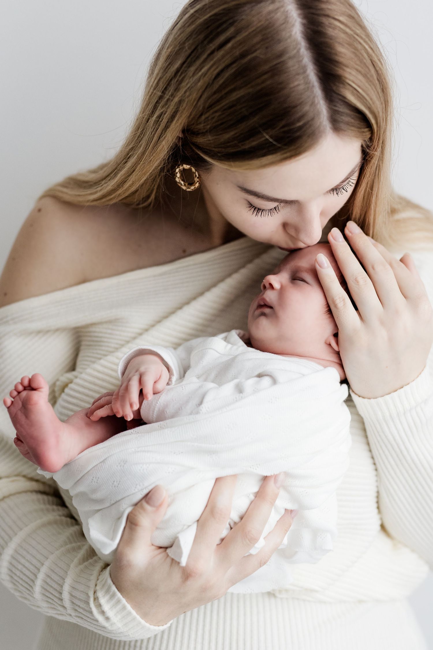 Young mother in white sweater gently kissing sleeping newborn baby wrapped in white cloth.