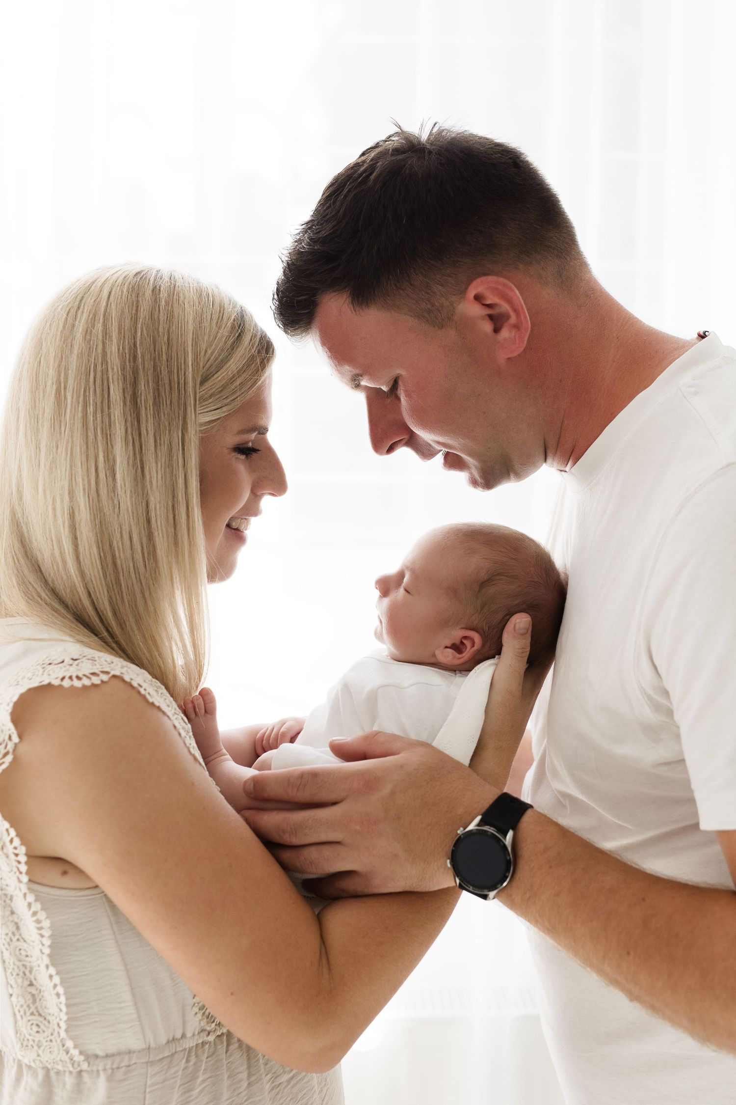 New parents lovingly gaze at their sleeping newborn baby held between them in a bright white studio.