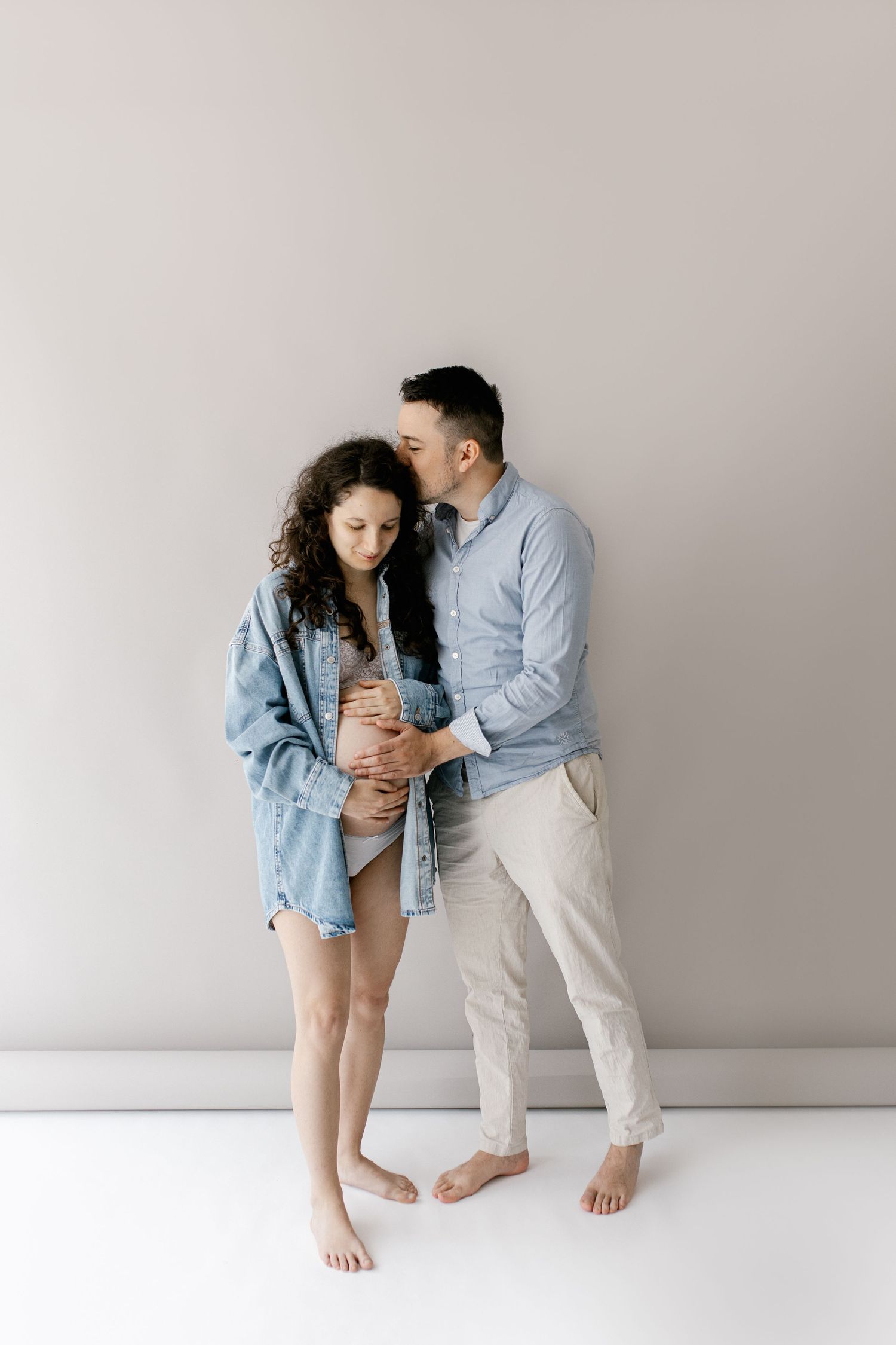 Couple expecting baby, man kissing pregnant woman's head while embracing her bump against gray wall.