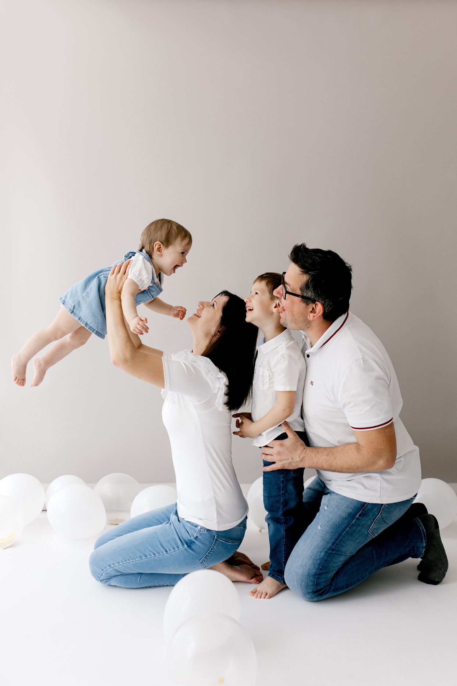 Happy family with two kids playing with white balloons on white floor, mom lifting baby girl while dad holds son.