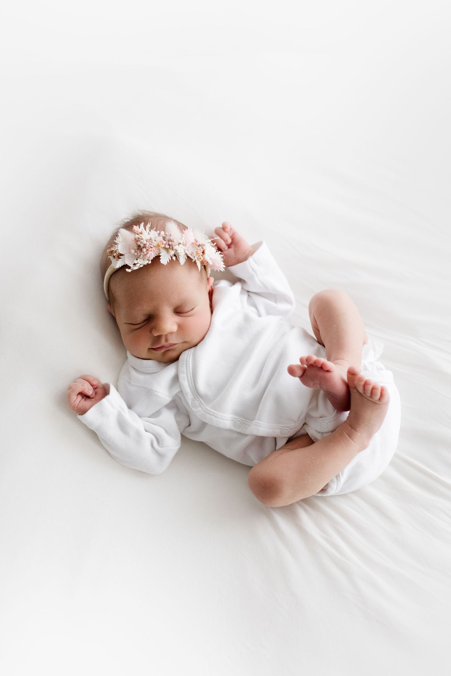 Newborn baby girl in white outfit and pink floral headband sleeping on white blanket.