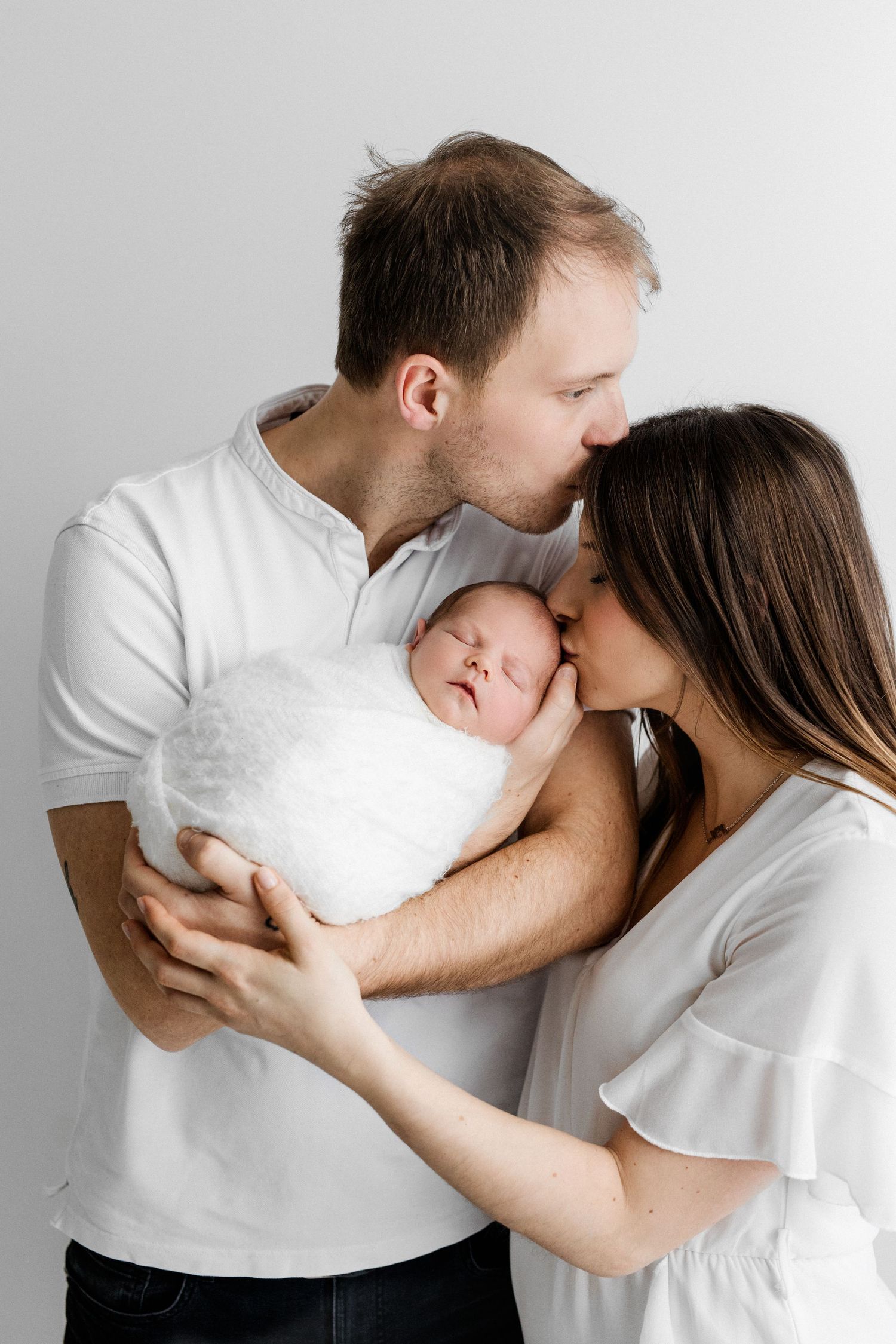 Happy parents kissing their sleeping newborn baby wrapped in white against a light background.