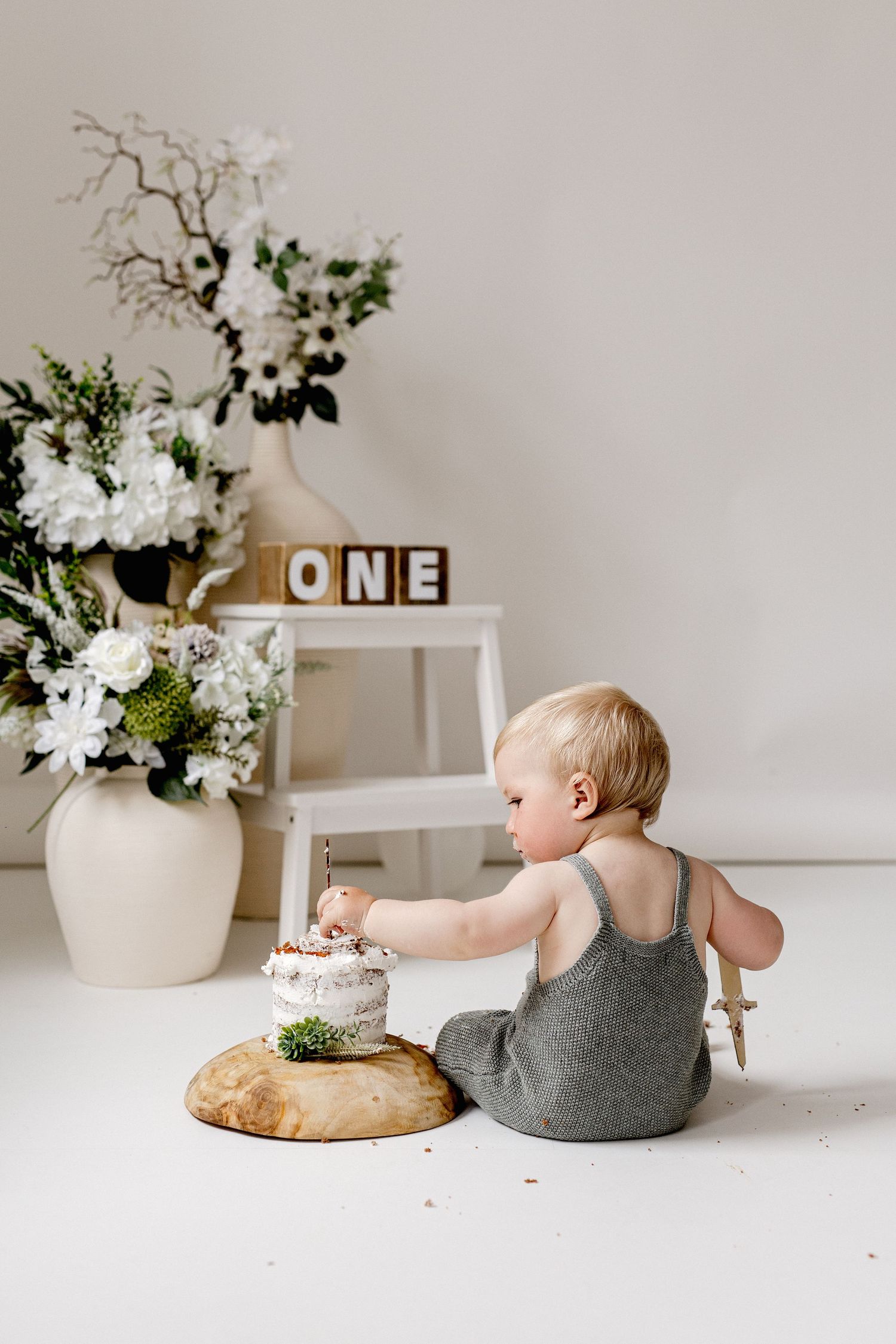 Baby in gray romper reaching into first birthday cake smash with white flowers and ONE blocks.