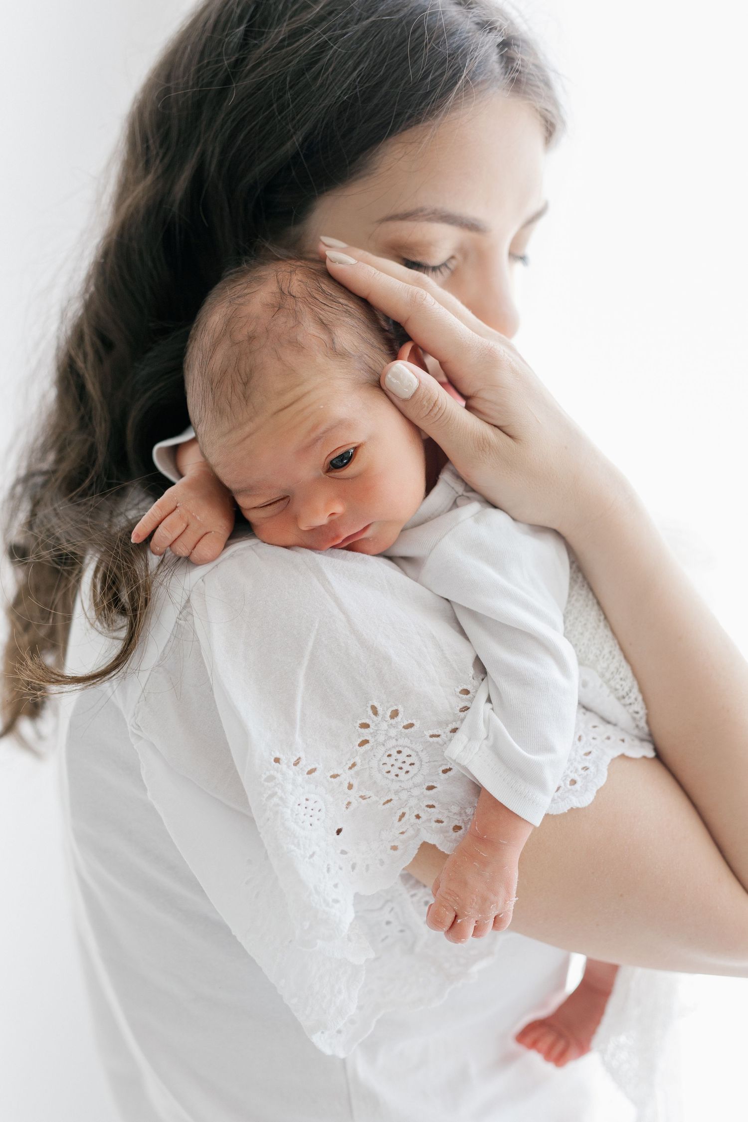 Tender moment of dark-haired mother in white lace dress cradling sleeping newborn baby.