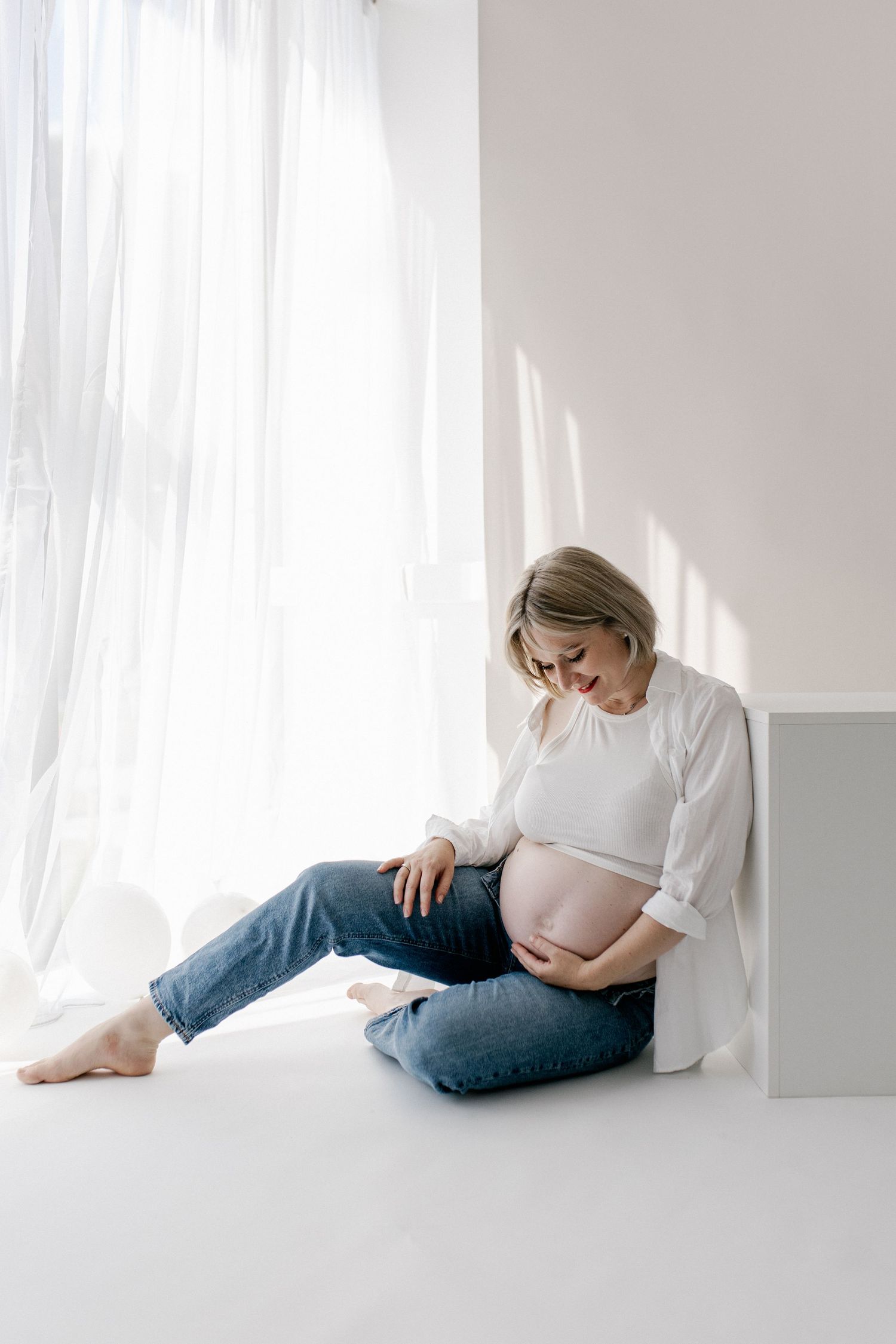 Pregnant woman in white top and jeans sitting on floor by window cradling her baby bump.