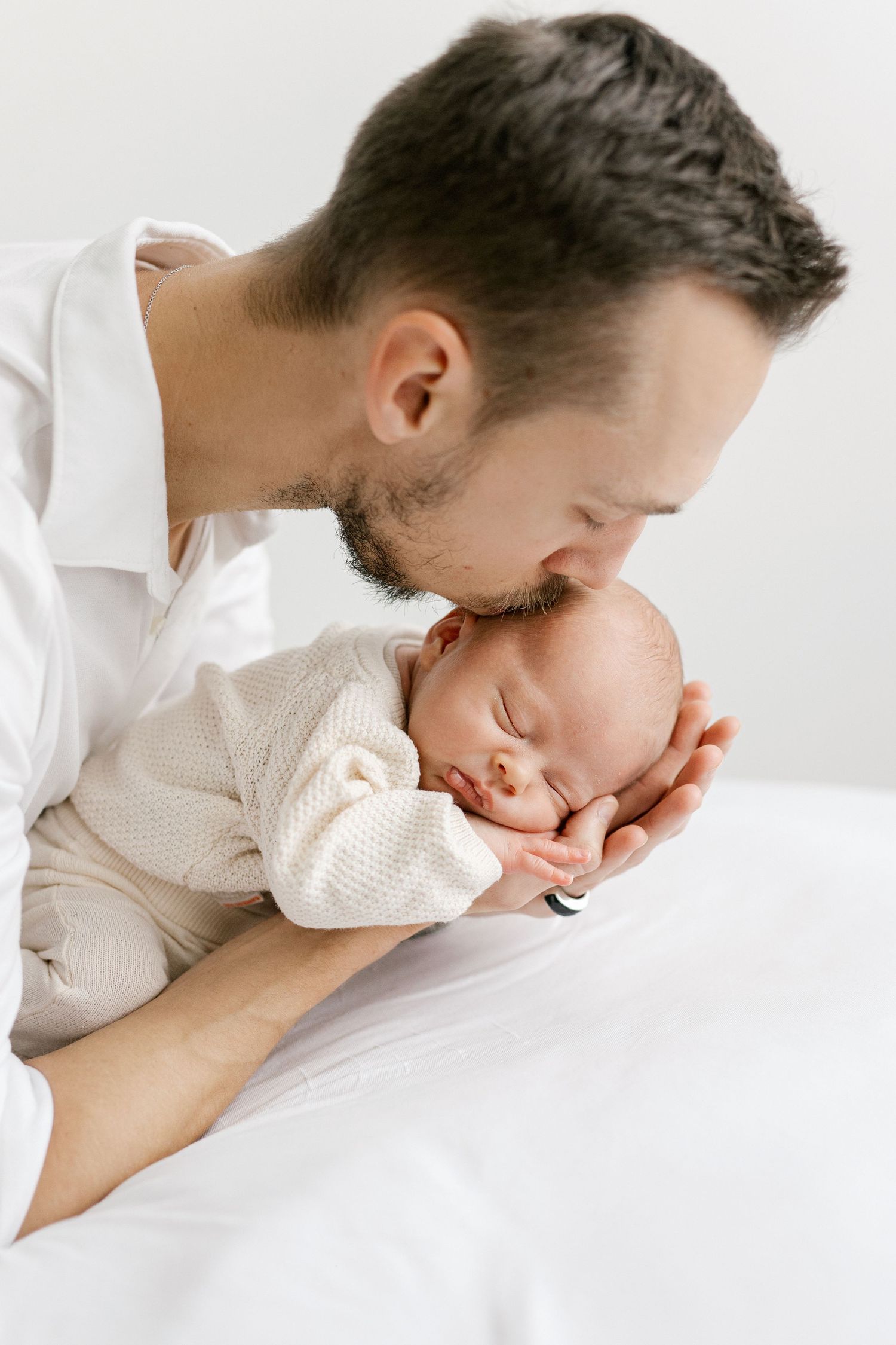 Father in white shirt gently kissing sleeping newborn baby cradled in his hands.