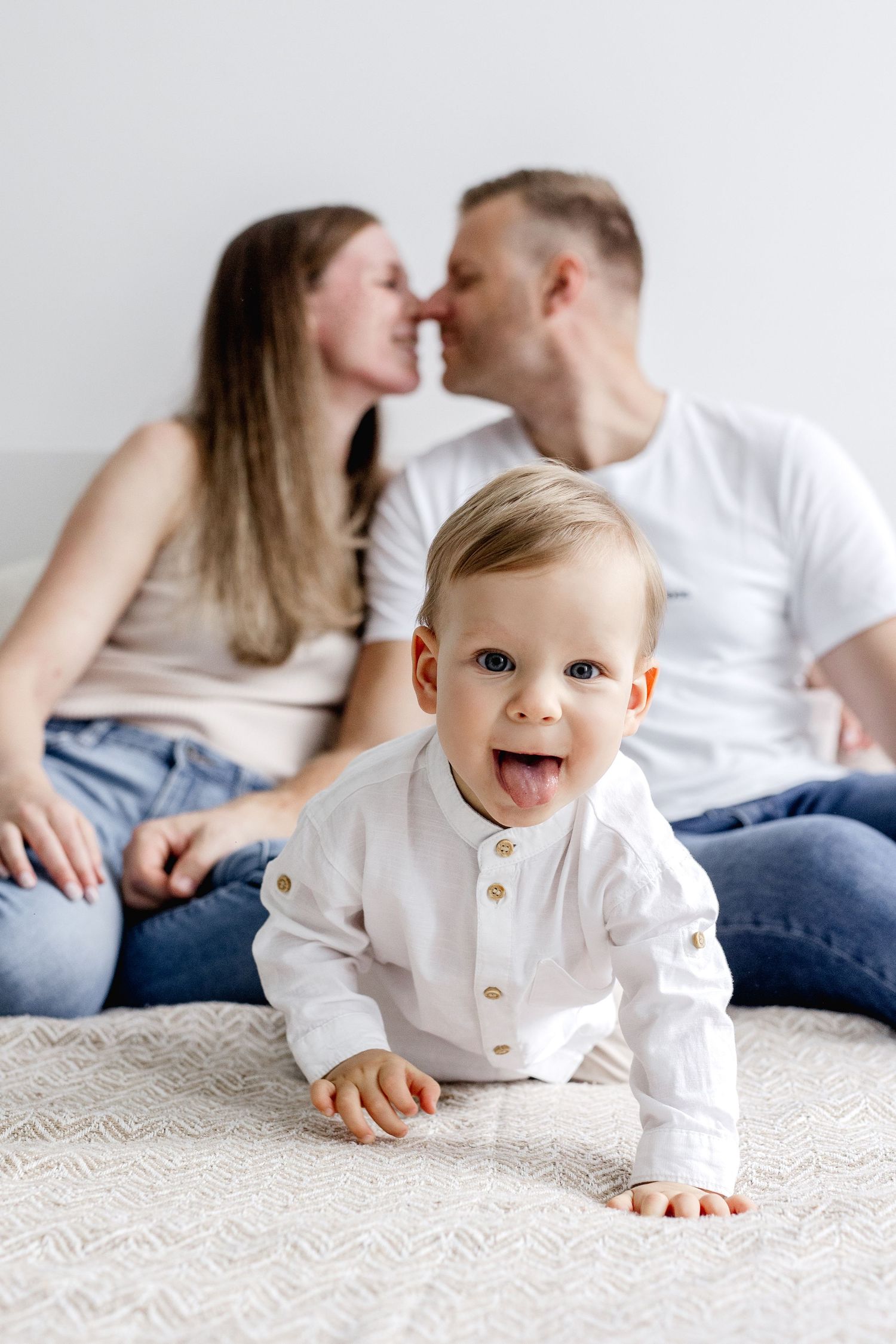 Happy baby in a white shirt crawling toward the camera while parents share a kiss in the background.