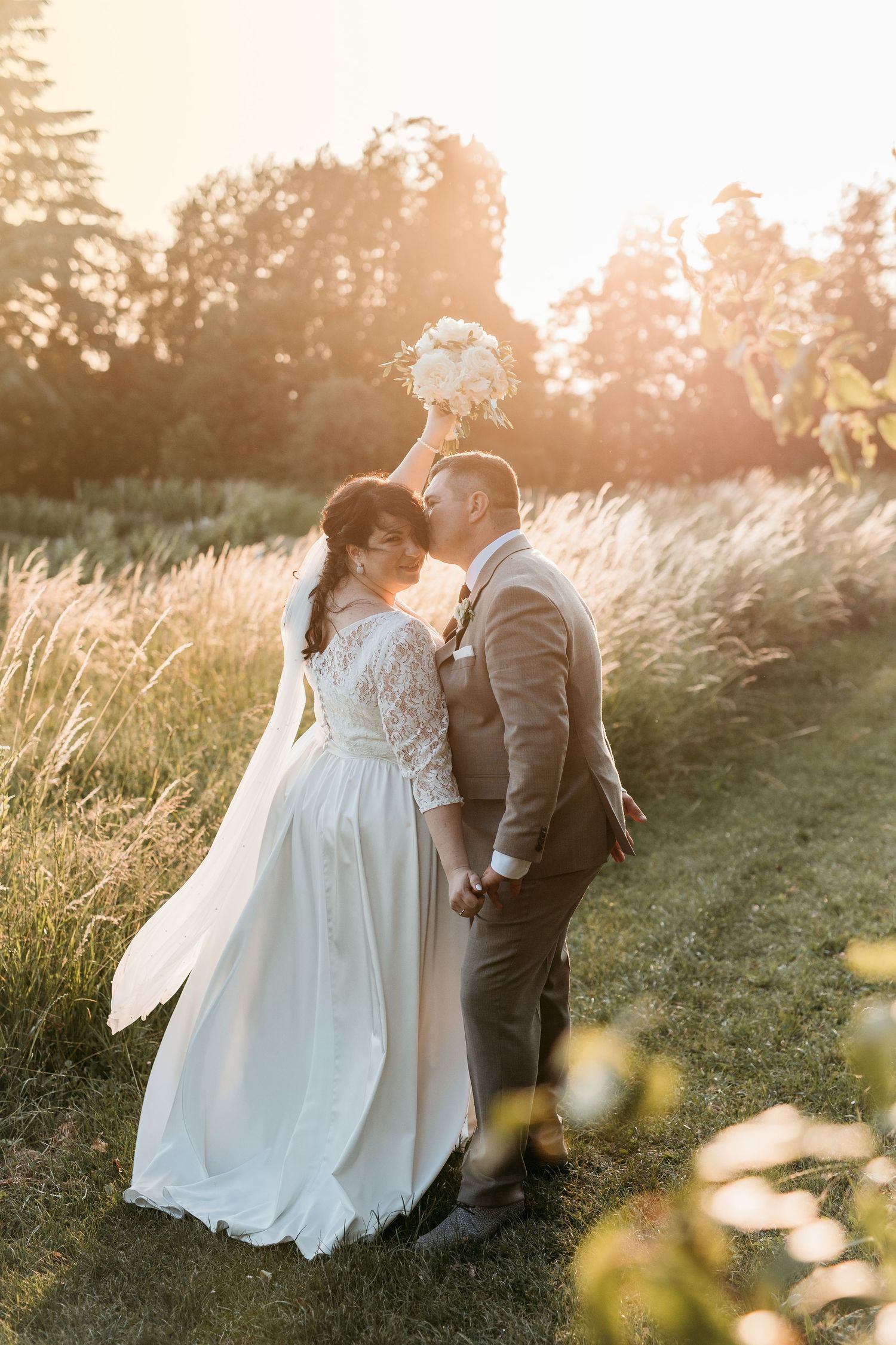 A bride and groom share a kiss at golden hour in a lush green field, bride holding a white bouquet overhead.