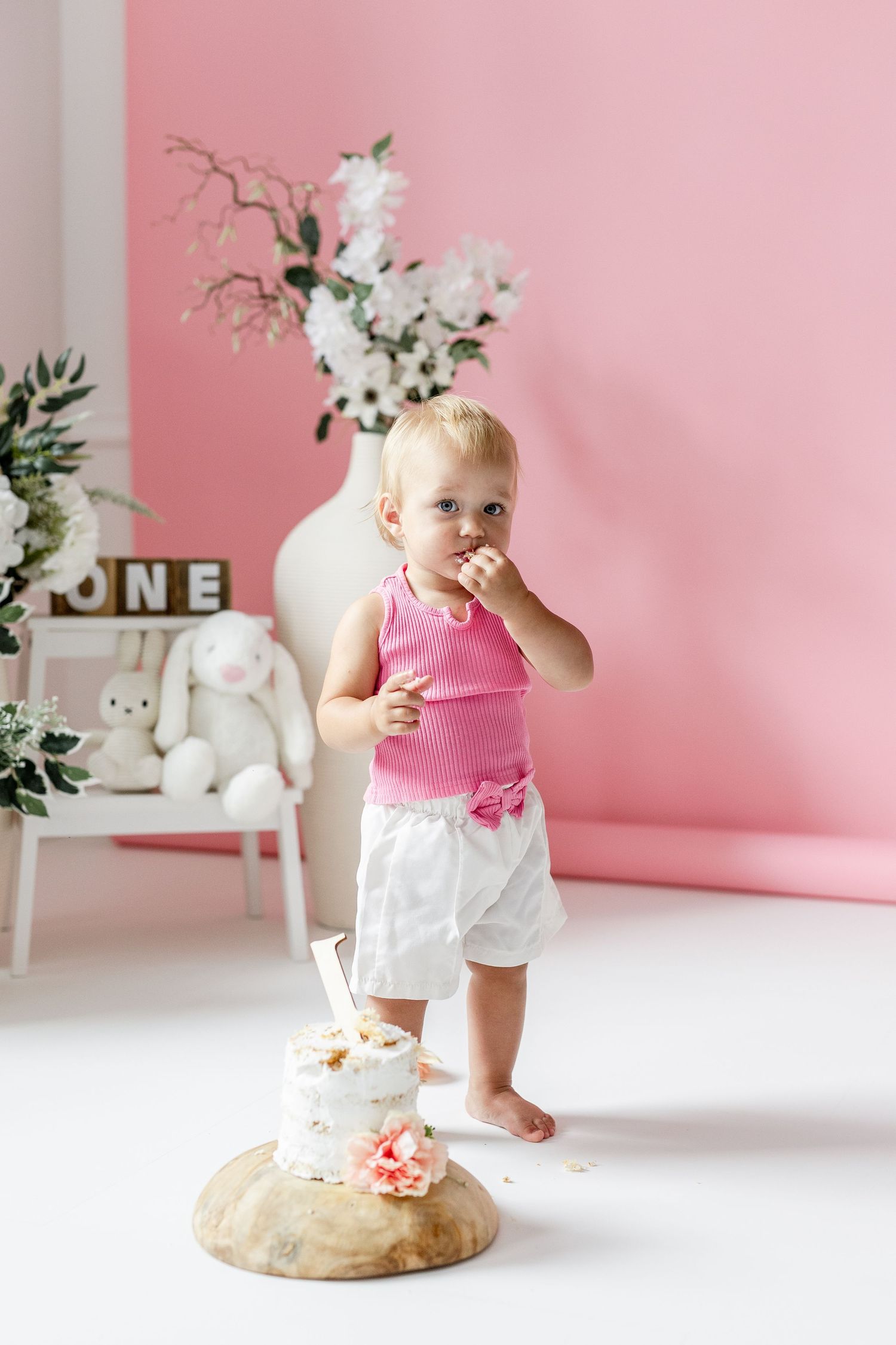 Baby girl in pink top and white shorts standing by smashed first birthday cake.