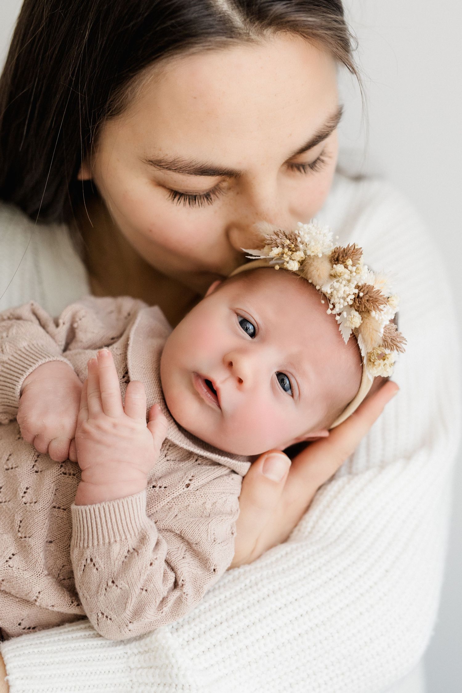 Dark-haired mother tenderly kissing her alert newborn baby girl wearing a floral headband.