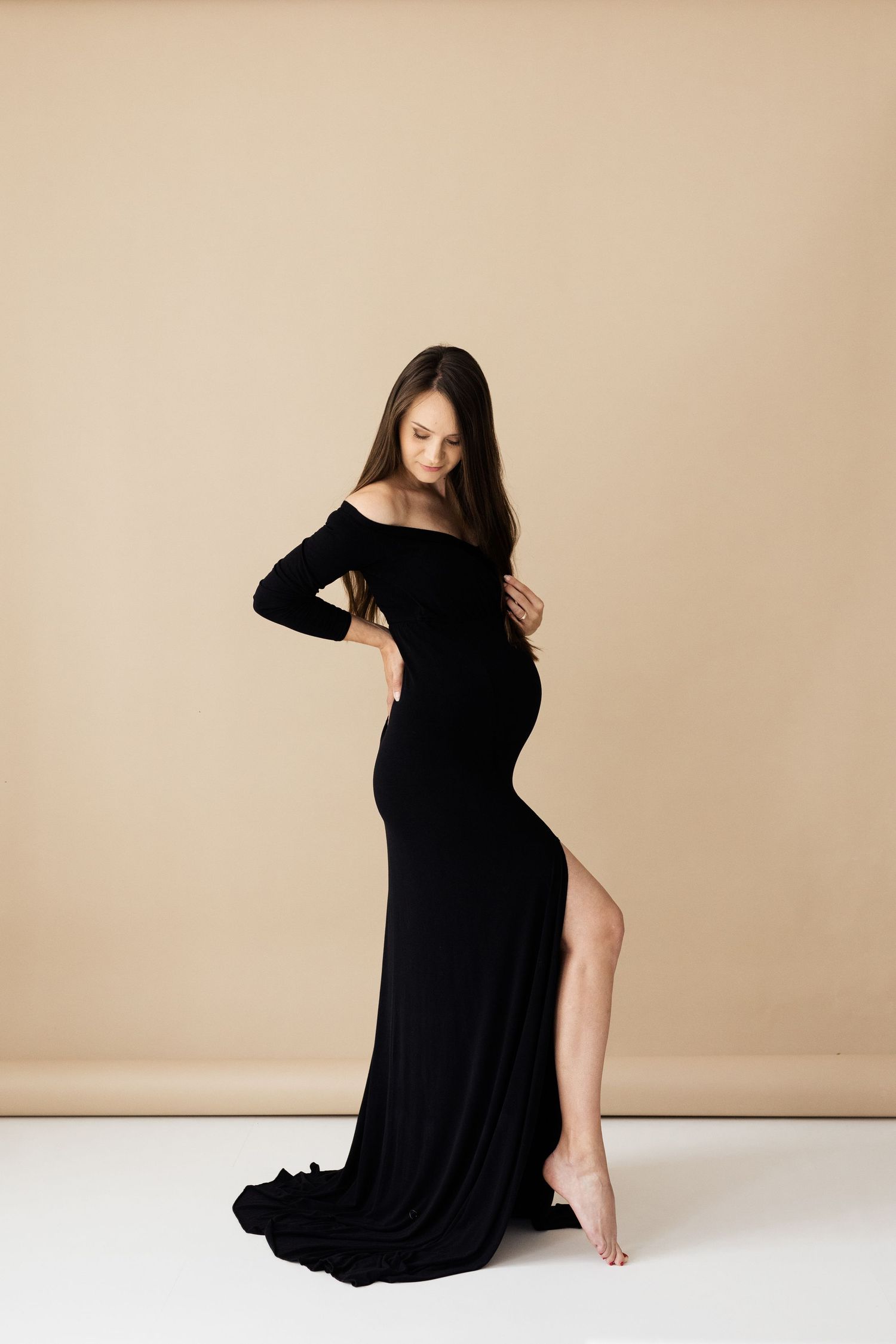 Elegant pregnant woman in black off-shoulder gown with slit posing in studio.