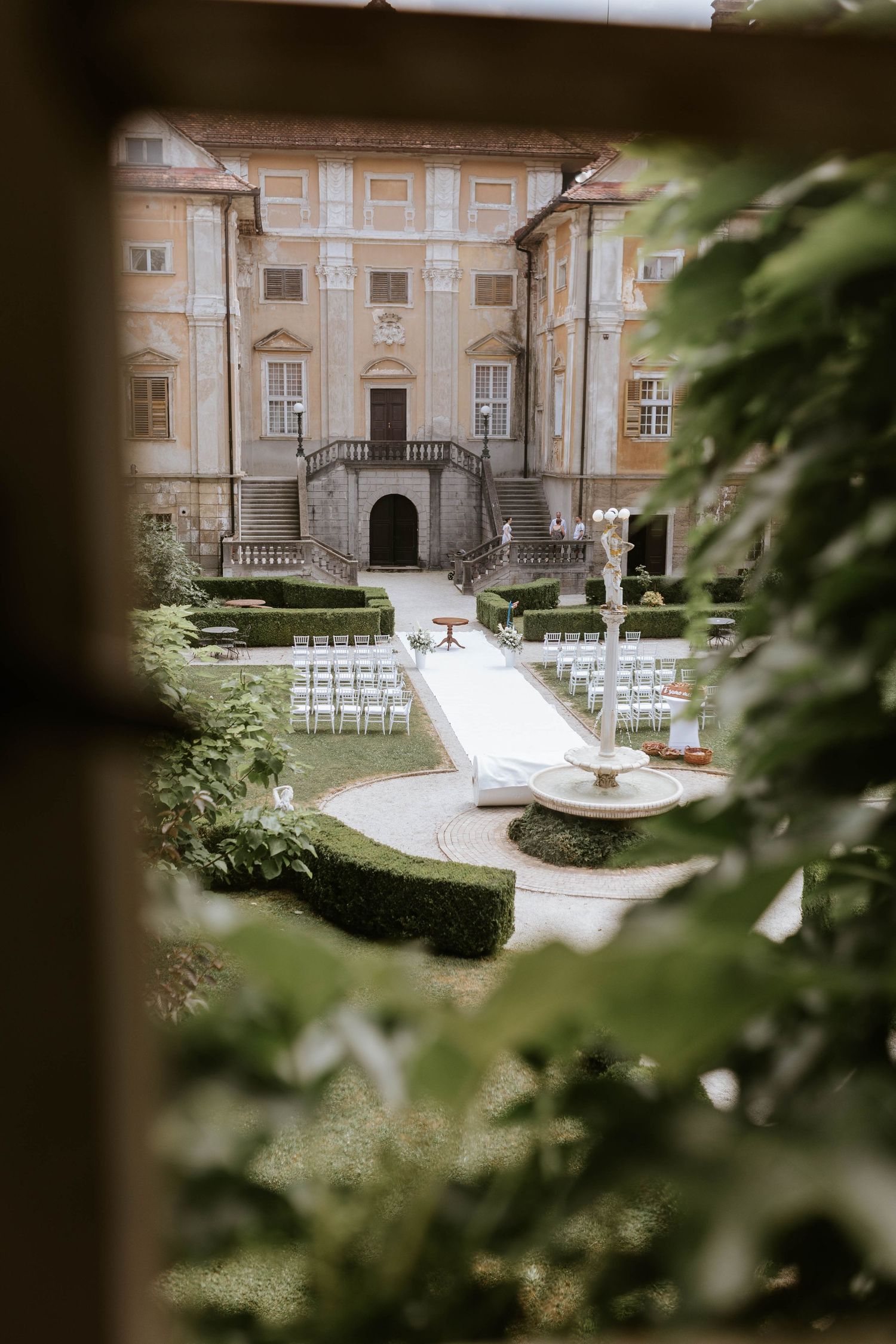 Baroque castle garden wedding ceremony setup with white chairs and fountain viewed through foliage.