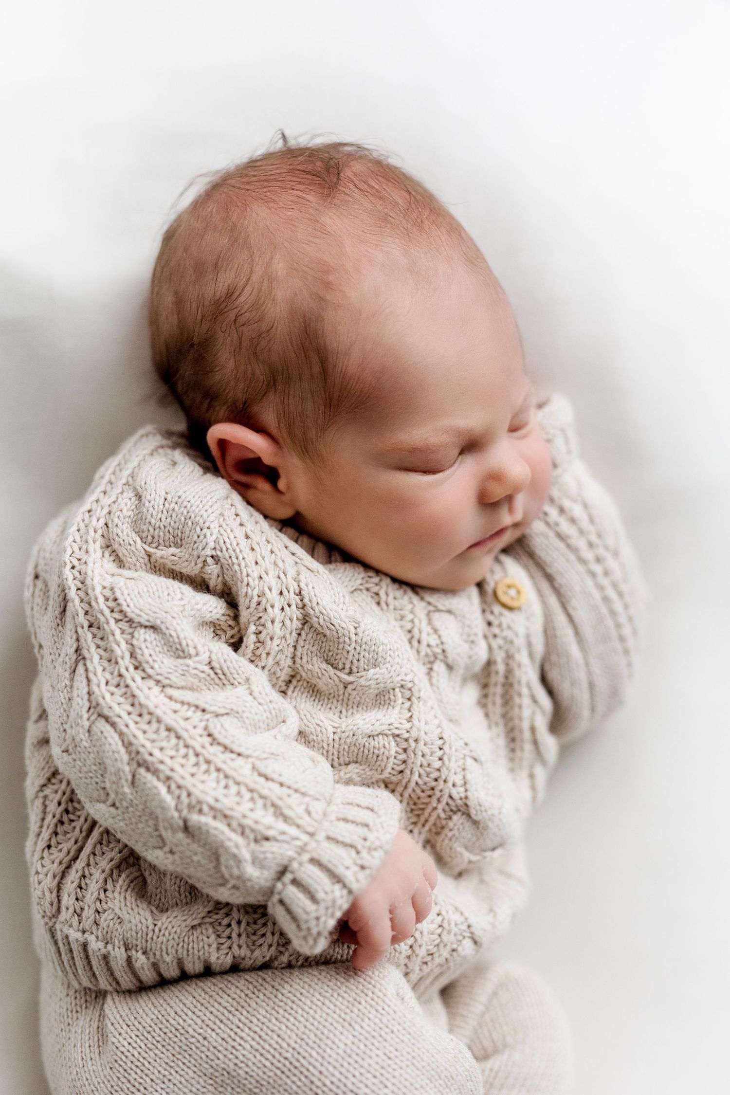 Close-up of sleeping newborn baby in beige cable-knit romper on white background.