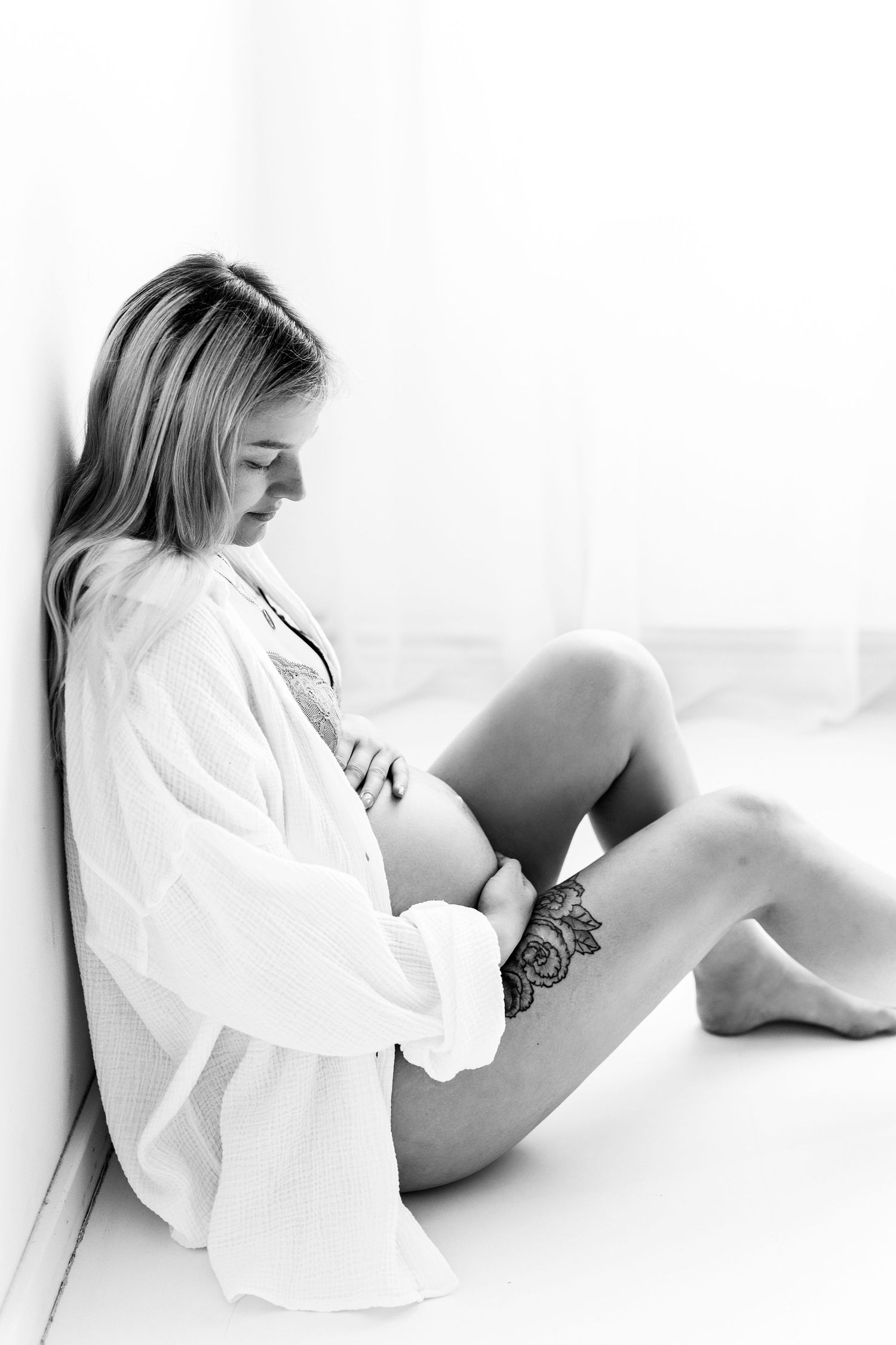Black and white photo of pregnant woman with tattoo sitting against wall in white shirt.
