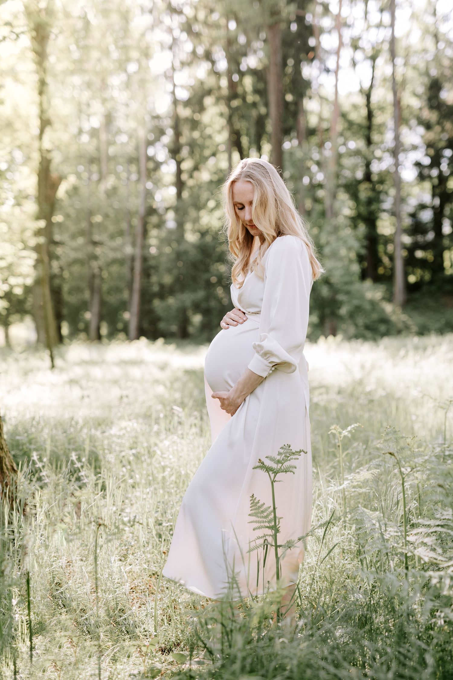 A pregnant woman in a flowing white dress cradles her baby bump in a sunlit forest meadow.
